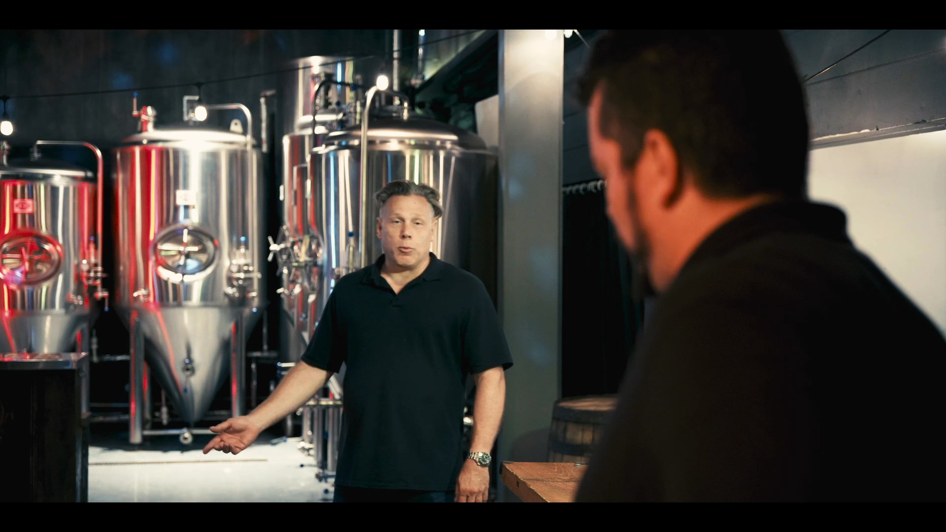Two professionals discuss operations within a commercial brewing facility, standing amidst rows of industrial stainless steel fermenting tanks.