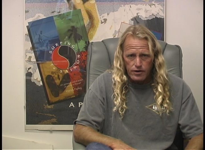 A man with long blonde hair sits in an office setting and engages in a serious, expressive discussion while gesturing to emphasize his points.