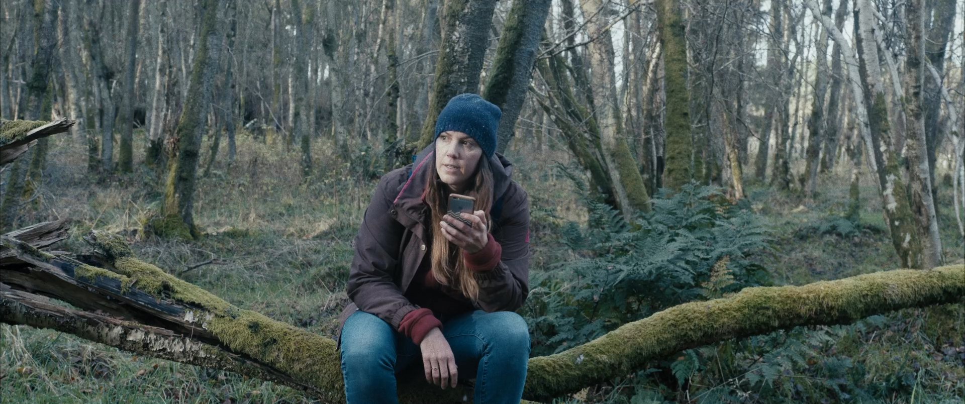 A woman sits on a mossy fallen tree in a forest, intently listening to audio recordings on her smartphone. She pauses frequently to contemplate the playback while surrounded by bare trees.