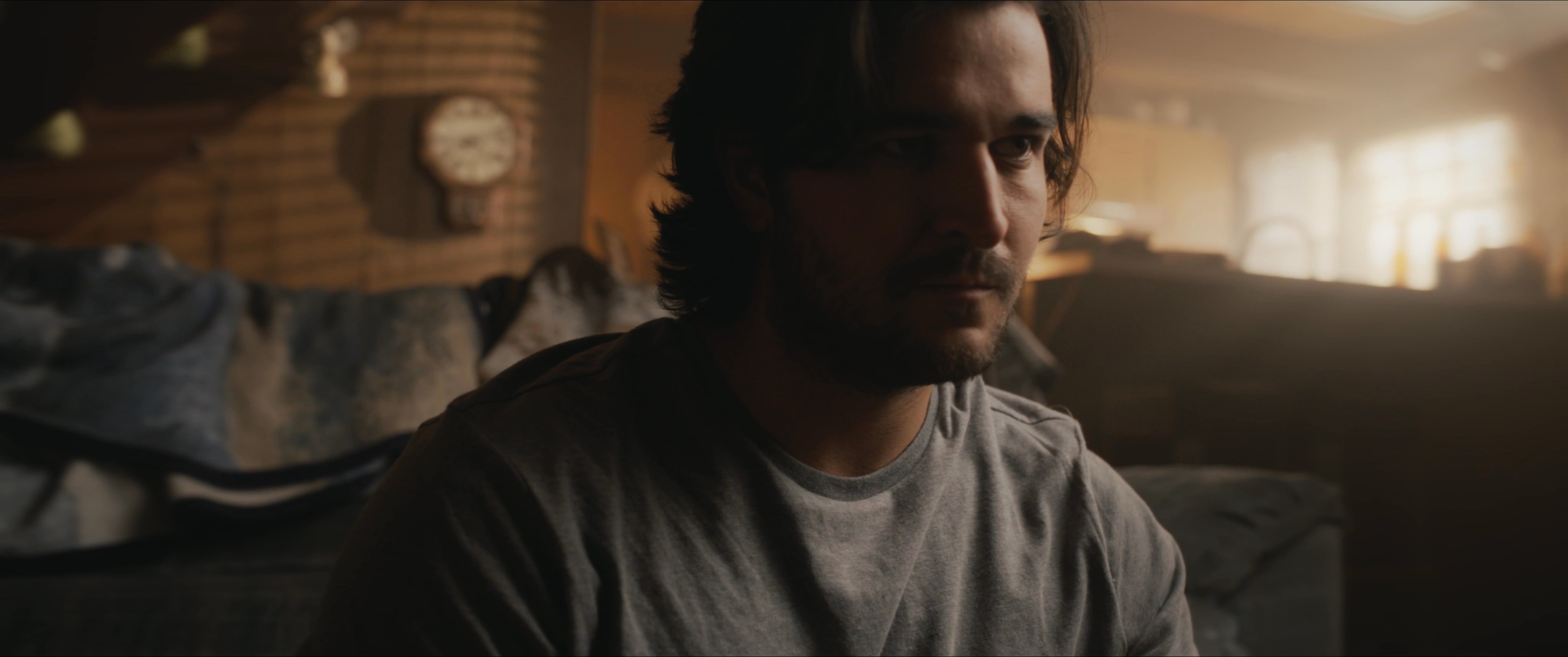 A man exhibits signs of anxiety and emotional distress while engaged in a tense phone conversation inside a dimly lit living room. The sequence concludes with him withdrawing into himself, reflecting a moment of significant personal burden.