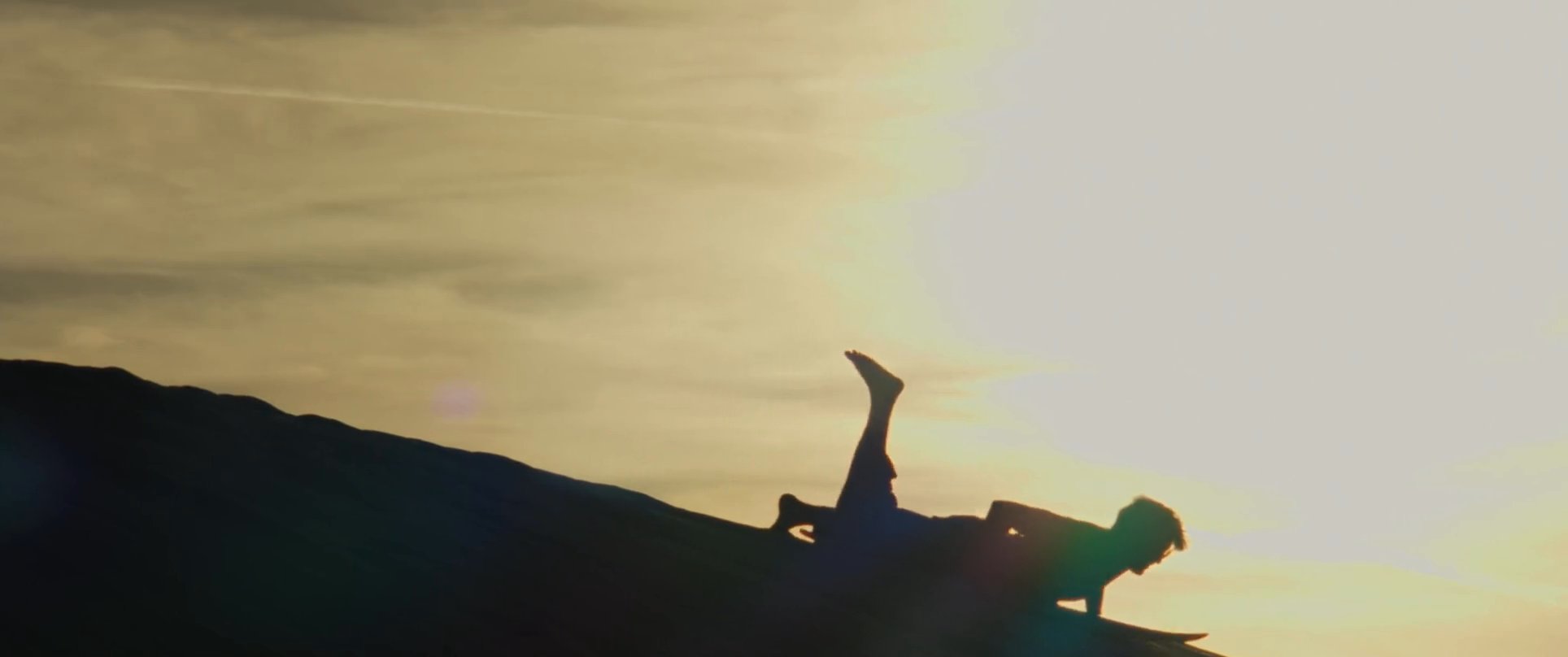 Two individuals stand with boards atop a vast dune at sunset before one descends the slope in a sandboarding run. The scene concludes with a wide landscape view of the desert horizon under a dramatic evening sky.