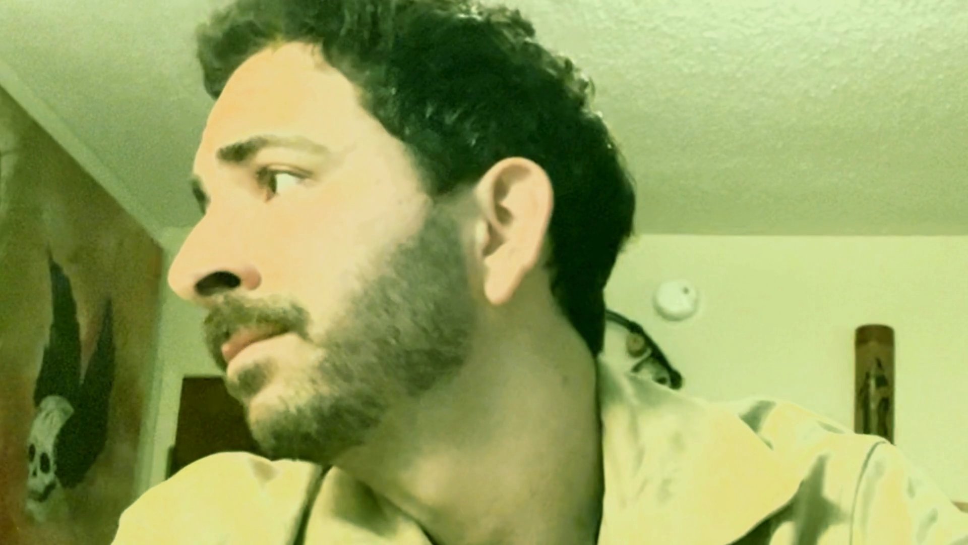 A man with a beard sits in an indoor space and speaks directly to the camera, sharing a message with expressive facial cues. The setting features decorative wall art in the background, framing the subject during his monologue.