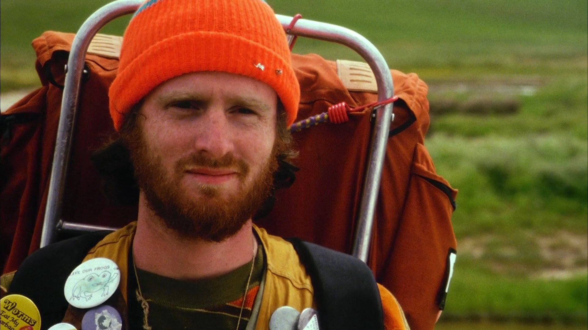 A woman in an orange hoodie interacts with a backpacker standing by a vintage car in an open field. The pair engages in a brief, poignant exchange before the hitchhiker kneels to gather belongings from the ground.
