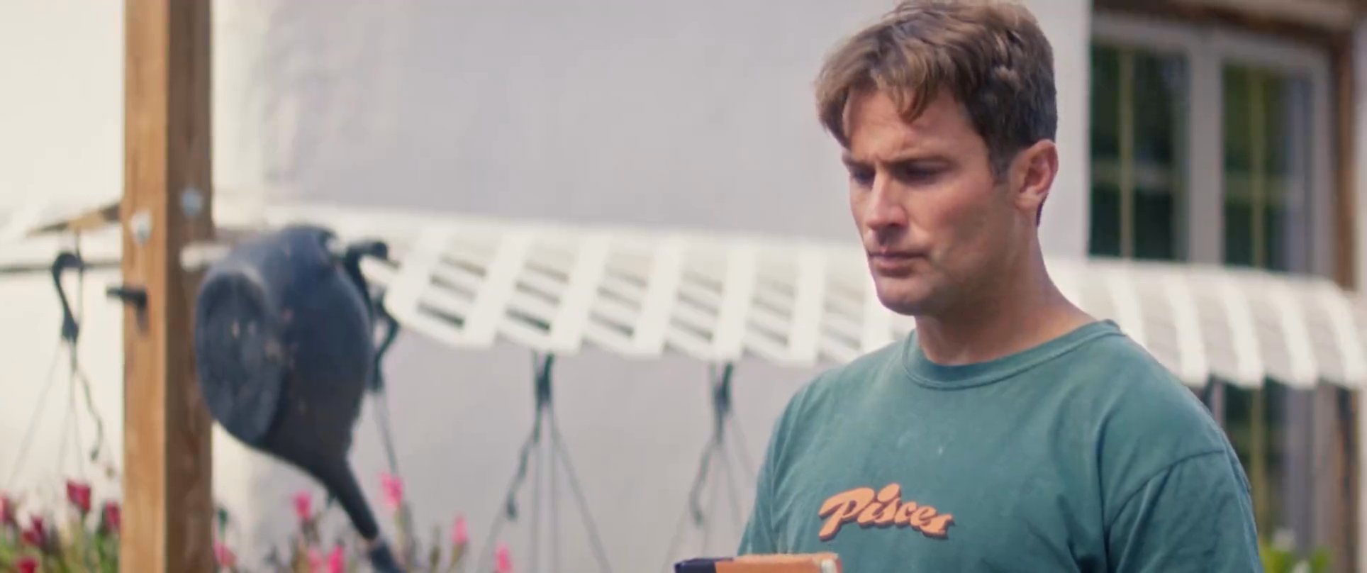 Two men engage in a serious conversation while standing on an outdoor patio, with one holding a book. The exchange features deliberate pacing and casual attire in a residential exterior setting.
