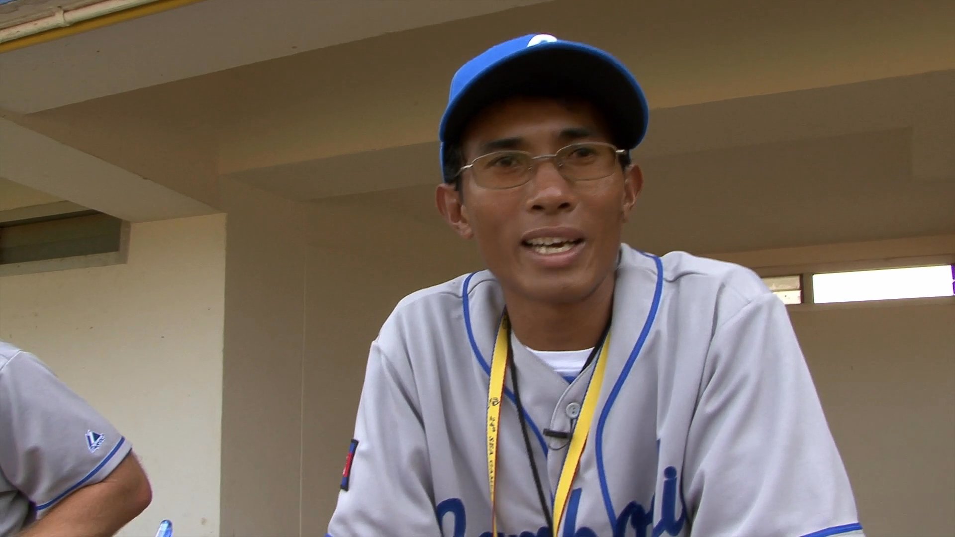 Baseball coaches in Cambodia national team uniforms discuss their training and program during an interview session at a stadium.