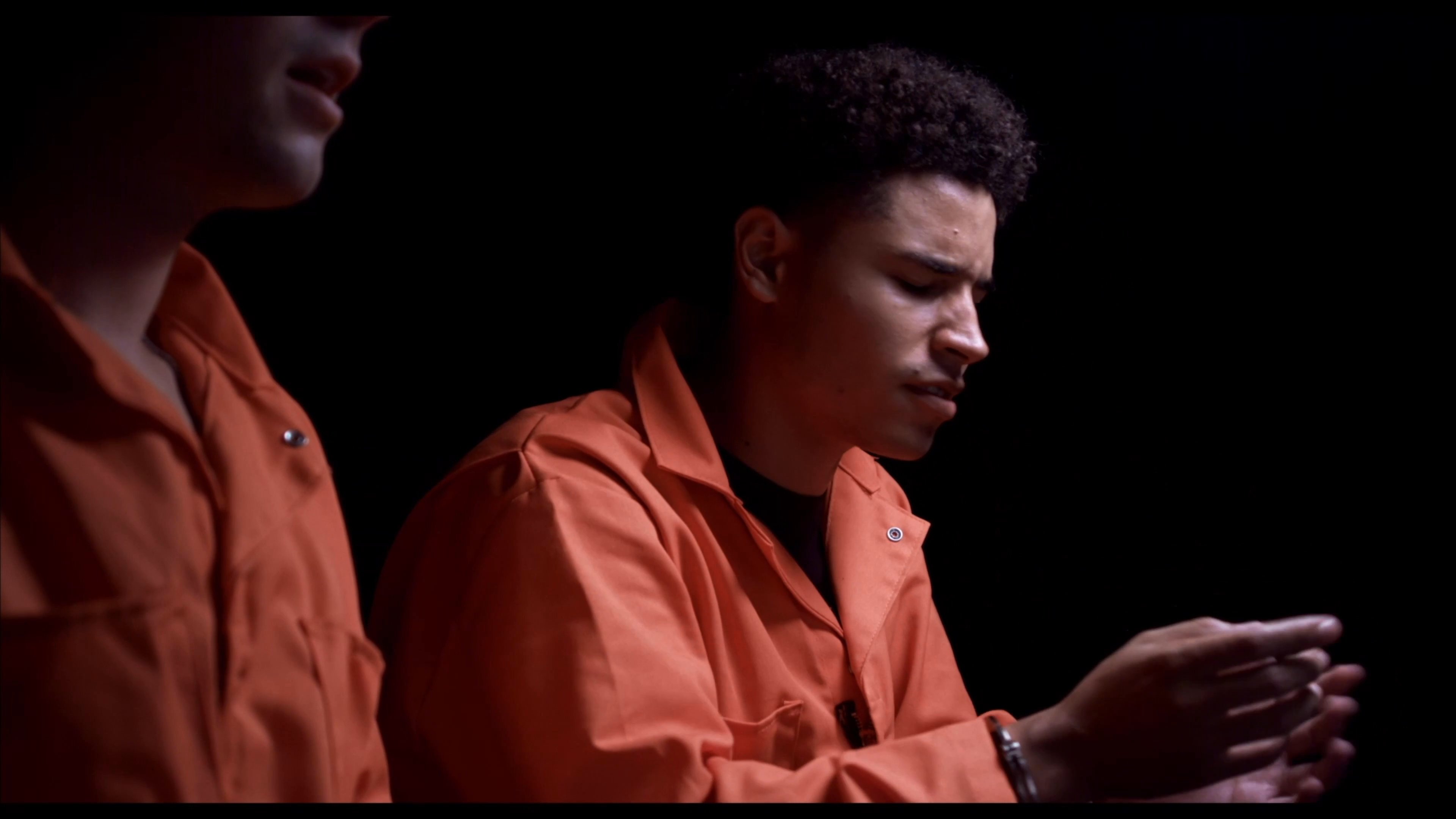 A DEA agent coordinates an investigation via radio while supervising an interrogation of a detainee wearing an orange prison jumpsuit. The scene alternates between the officer's tactical updates and the intensive questioning of the suspect in a dimly lit room.