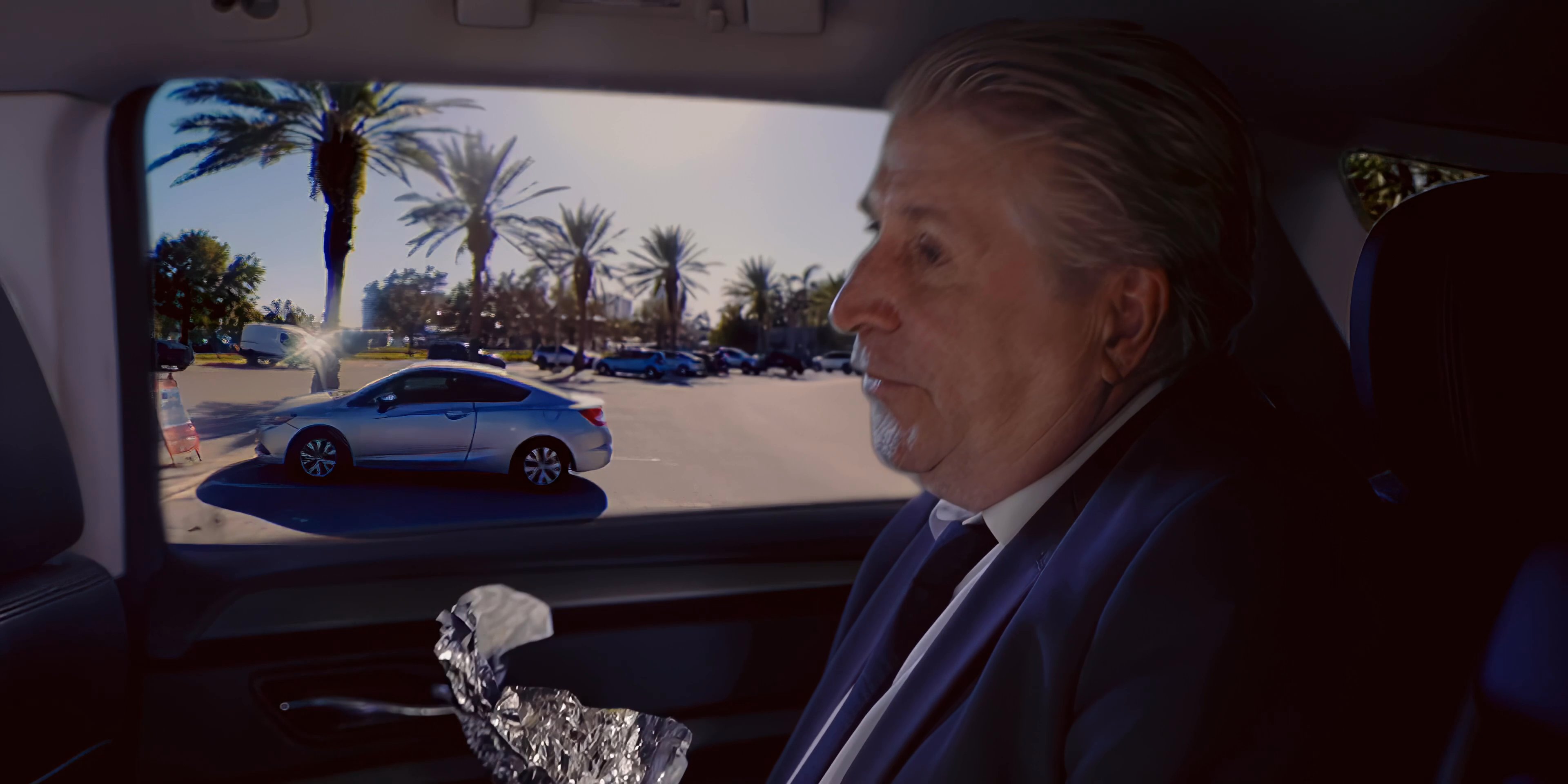 Two men hold a discussion while seated in the back of a vehicle, with one individual eating a wrapped meal during the exchange.