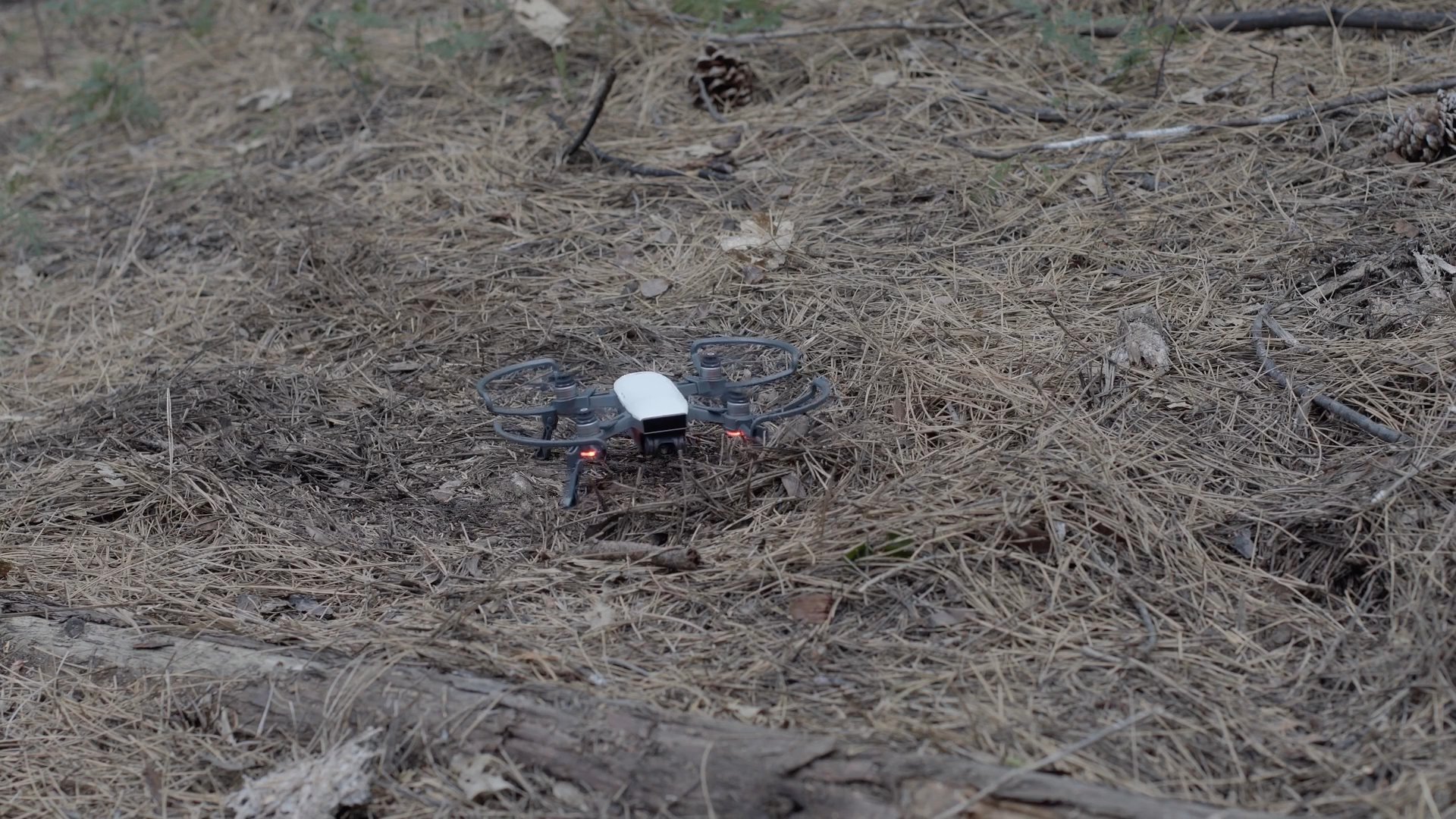 An injured woman lies on the ground in a wooded area while a small drone navigates the environment to locate her. The aerial device maneuvers around tree trunks and stumps to assess her condition.