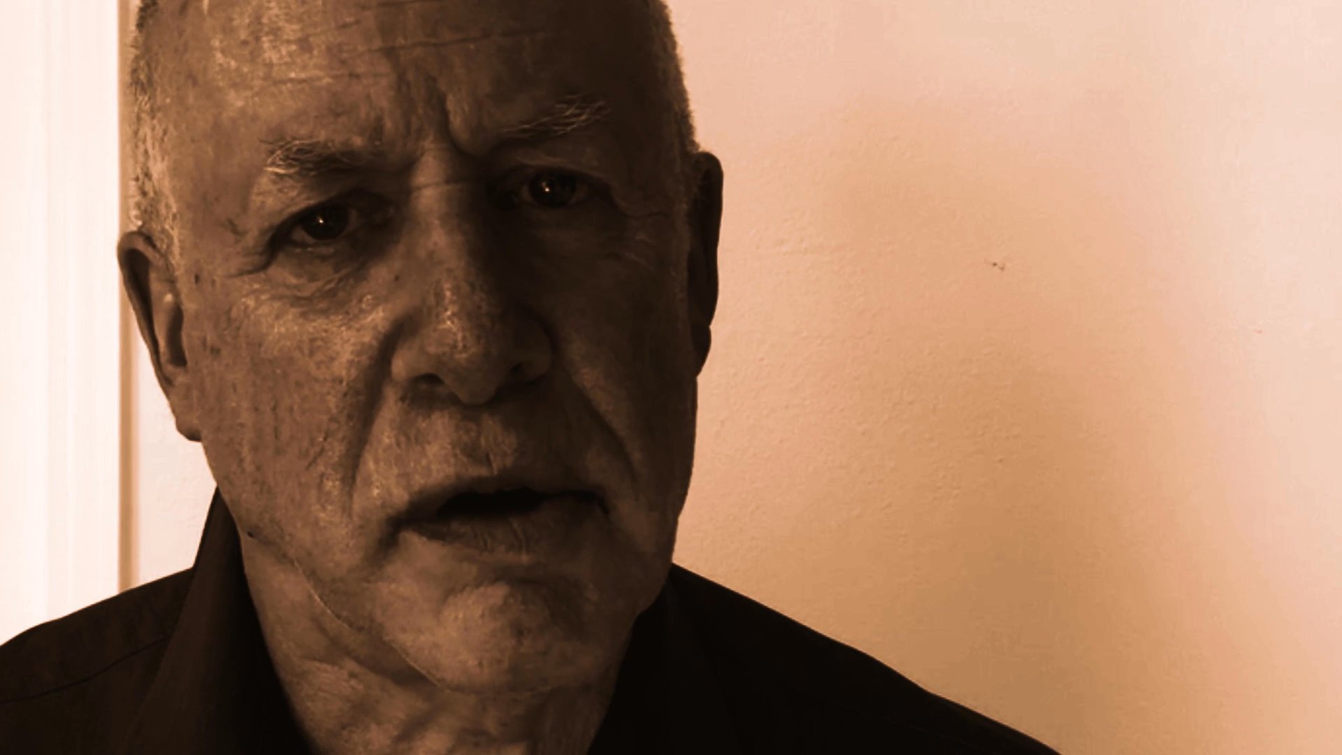 An older man faces the camera and speaks directly to the viewer in a sepia-toned environment. His steady gaze and measured delivery suggest he is sharing a personal testimony or narrative.