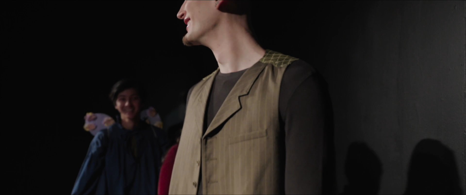 A group of actors stands on stage receiving applause from an unseen audience following a performance. The cast members smile while acknowledging the crowd in a dimly lit theater setting.