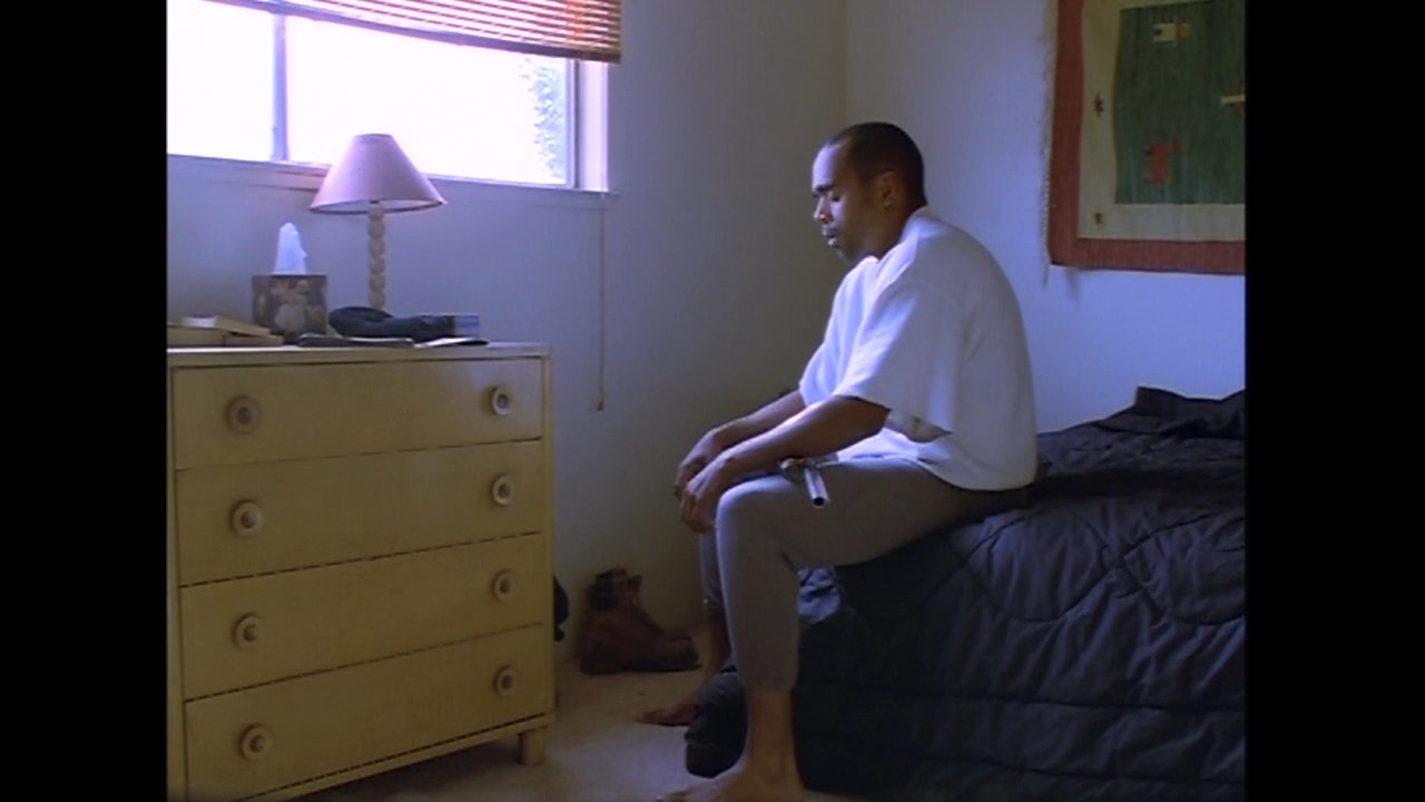 A man sits alone on a bed in a sunlit bedroom, introspectively holding a rifle in a moment of quiet gravity. The scene transitions from an exterior view of a suburban residence to the interior emotional landscape of its occupant.