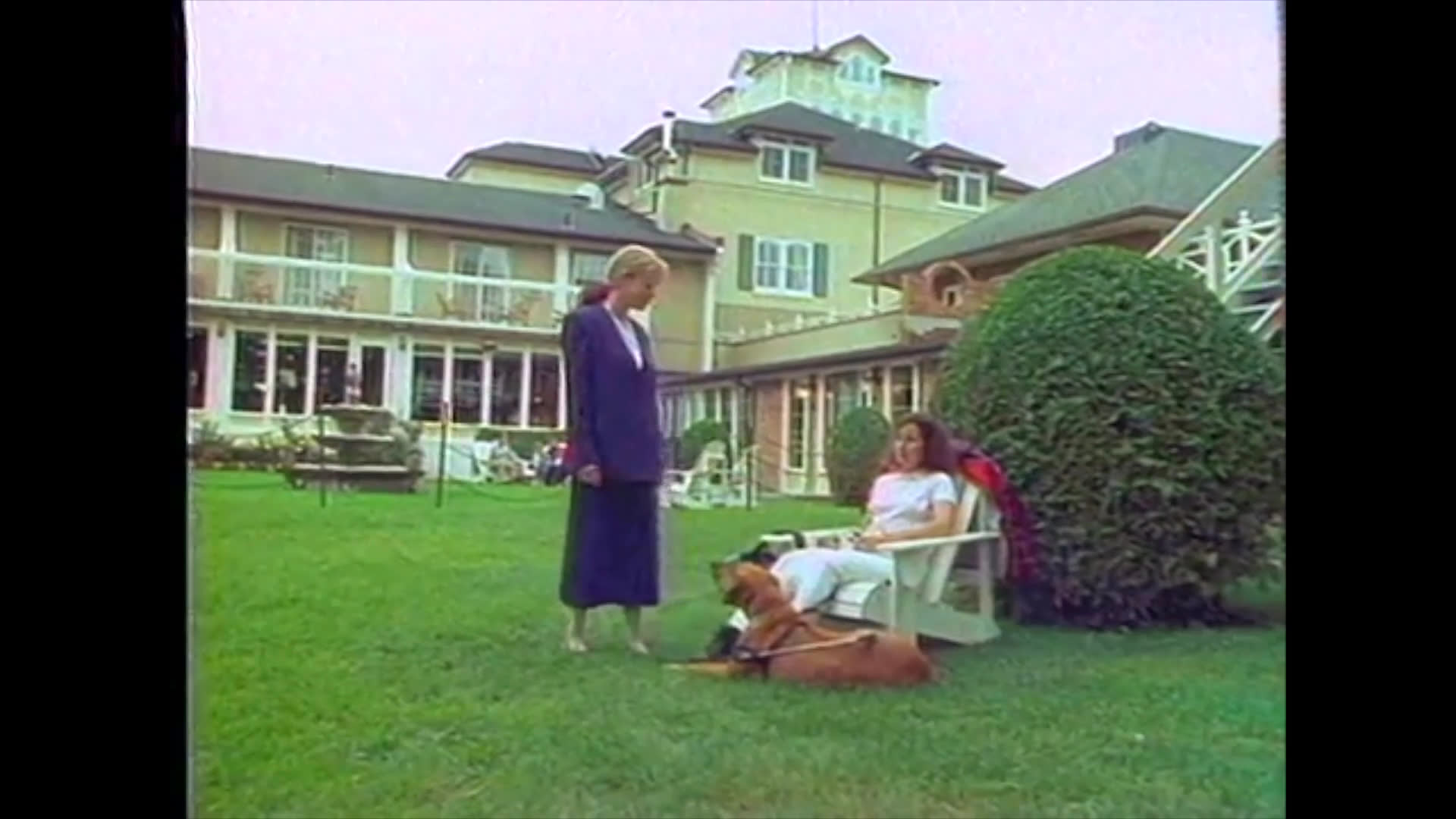 A woman accompanied by her service dog rests in a resort garden before being approached by another individual for a professional interaction. The setting transitions from a casual outdoor moment to a direct conversation between the two women.