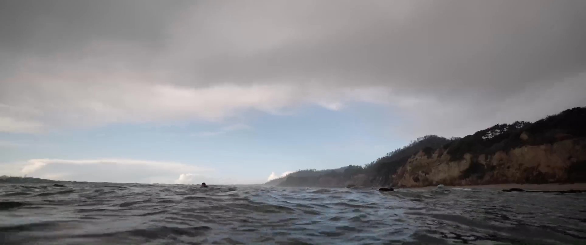 A surfer in a wetsuit maneuvers a surfboard through choppy ocean waves near a rocky coastline. The perspective shifts between underwater views and surface-level shots of the breaking surf.