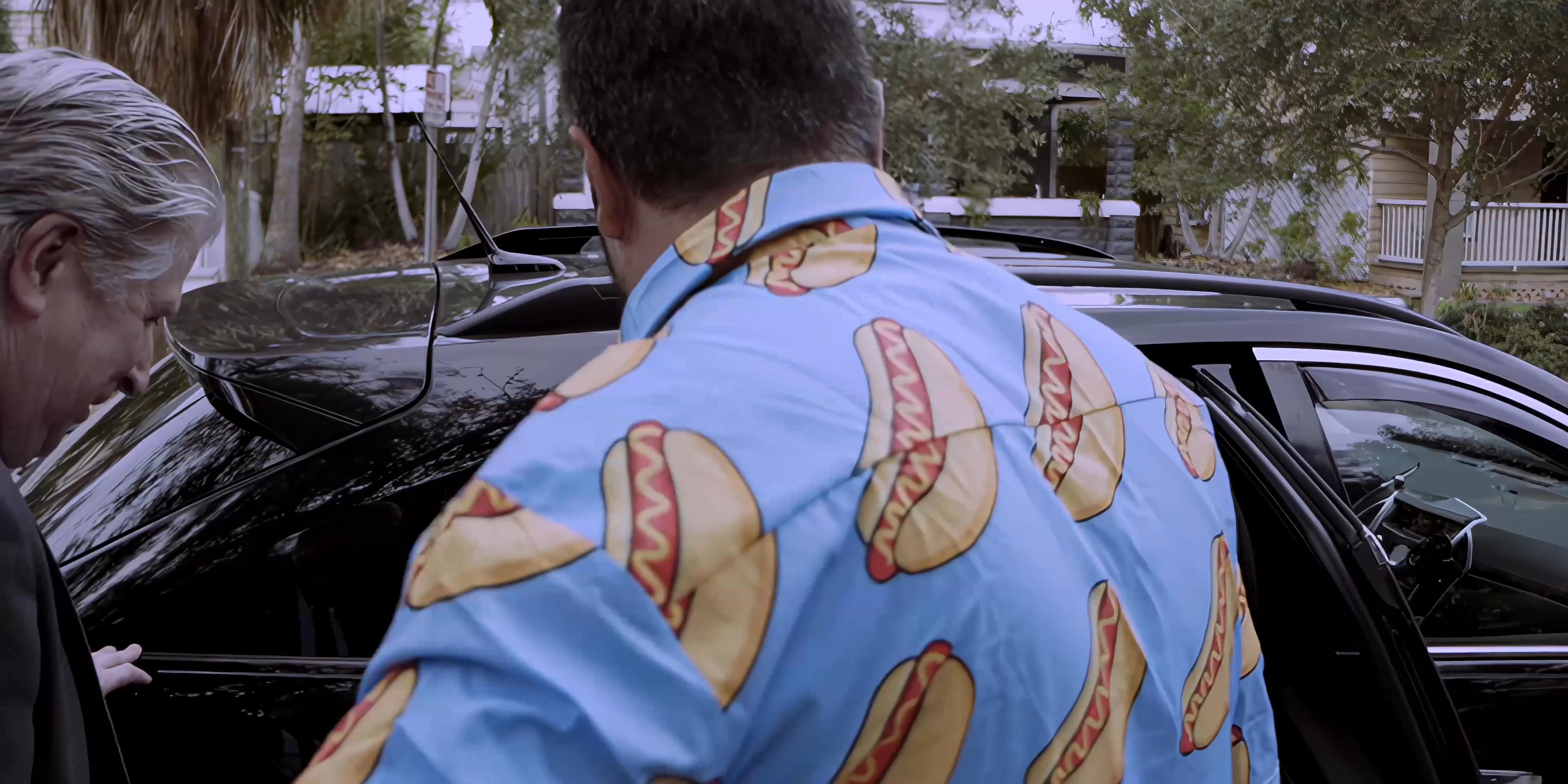 An older professional in a suit and a younger man in a vibrant hot dog print shirt engage in a brief outdoor conversation before boarding a vehicle together. The two men then sit in the back seat and continue their interaction as the car prepares for travel.
