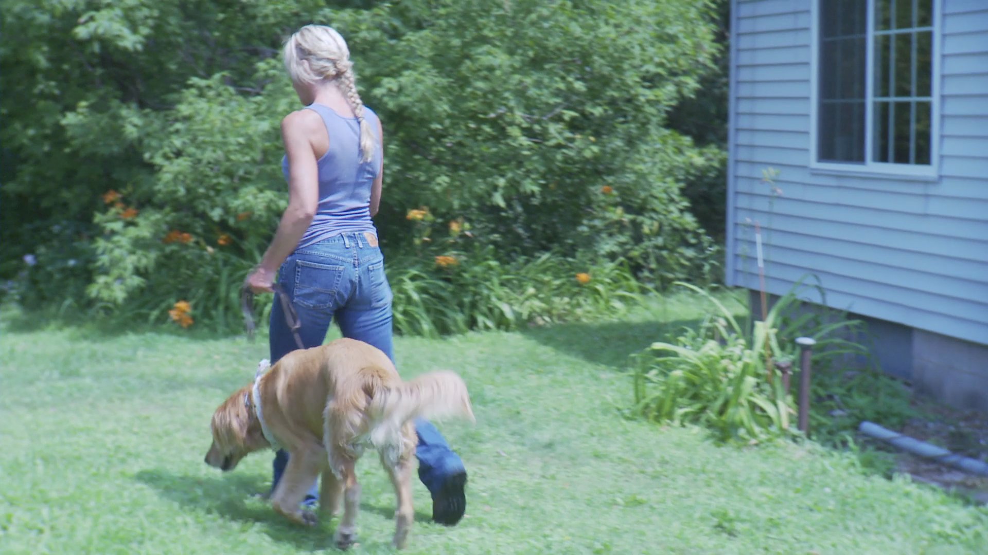 A woman prepares to leave her front porch and walks her golden retriever through a residential yard into a wooded area.