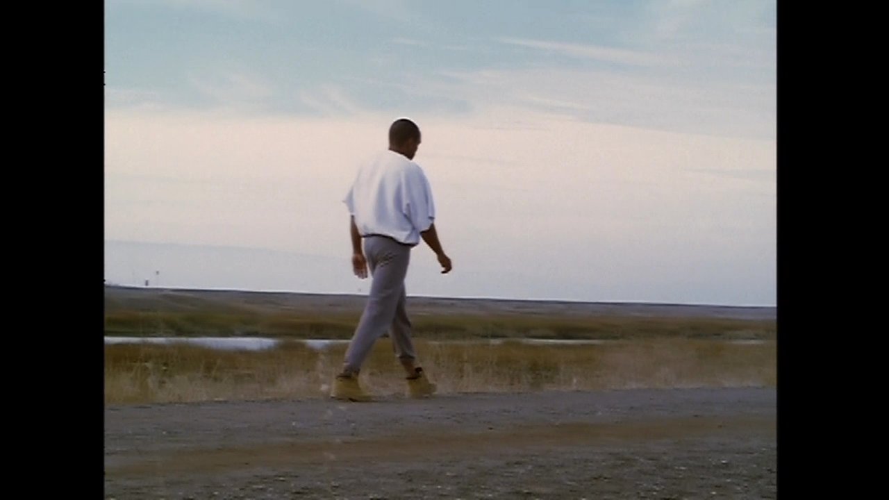 A man walks alone along a remote, rural roadside against a backdrop of distant hills, eventually transitioning into an urban setting.