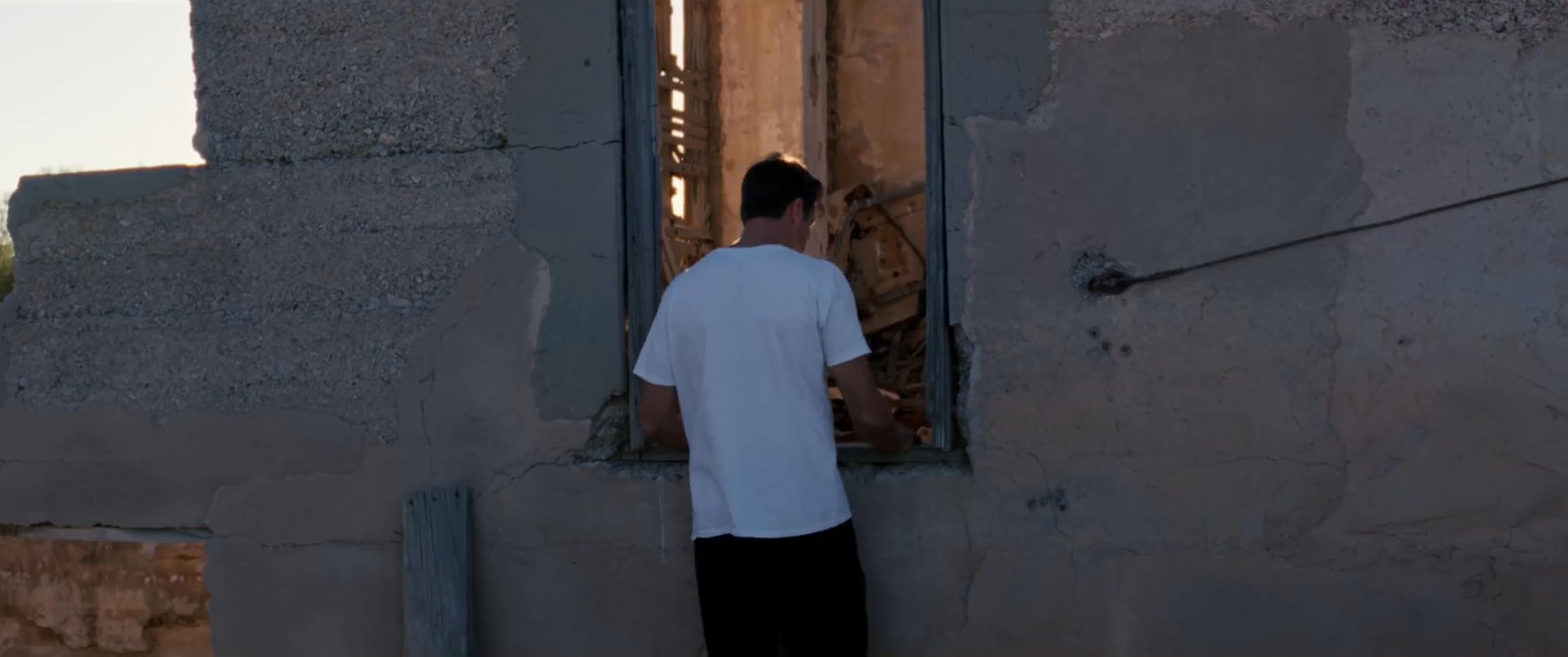 A man explores an abandoned, decaying building in an arid desert landscape as the sun sets. The scene transitions from the crumbling structure to a peaceful, darkening horizon.