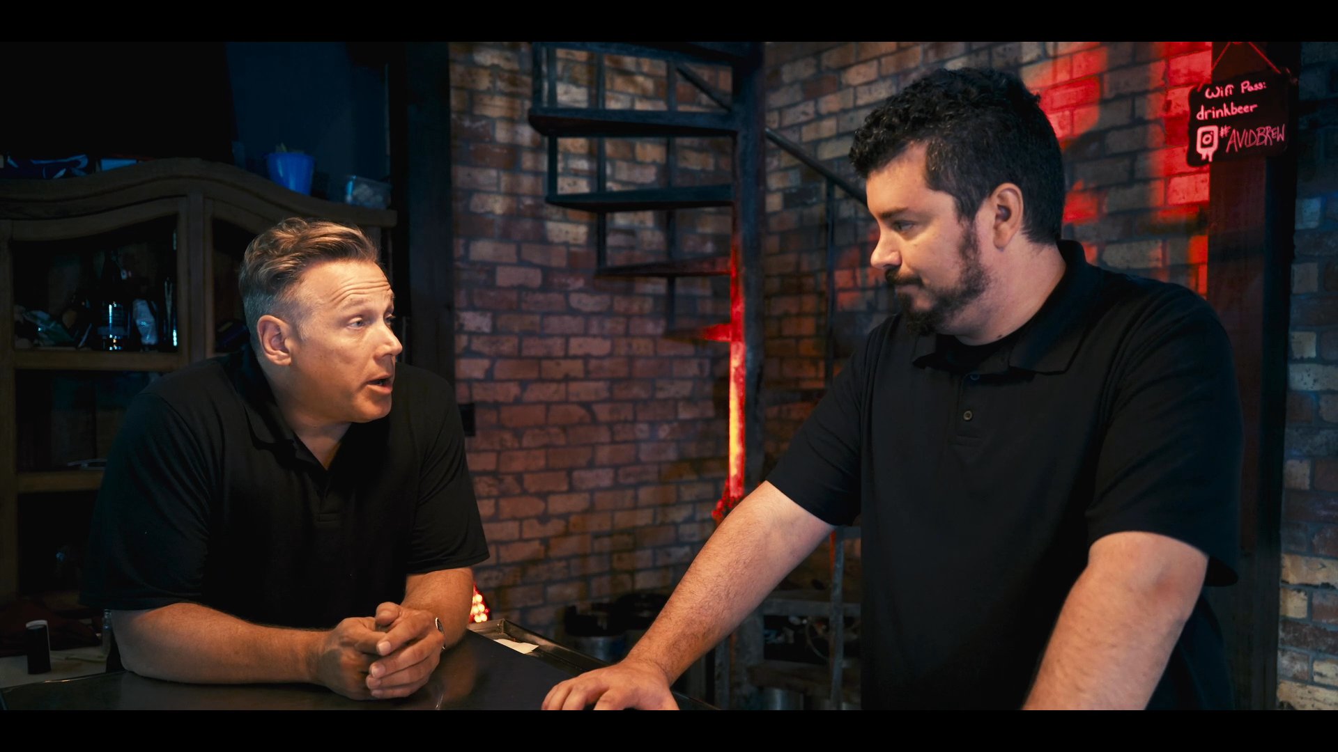 Two individuals engage in a professional discussion within a brewery setting, appearing to coordinate operations or business strategies. They maintain a focused, collaborative interaction while standing at the bar counter.