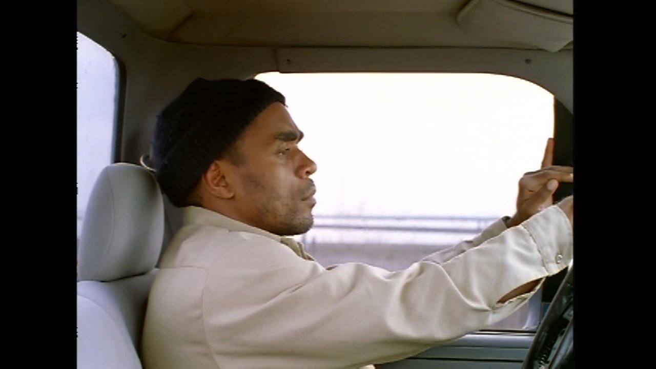 A man drives a red pickup truck along a dusty, rural road in a desert environment.