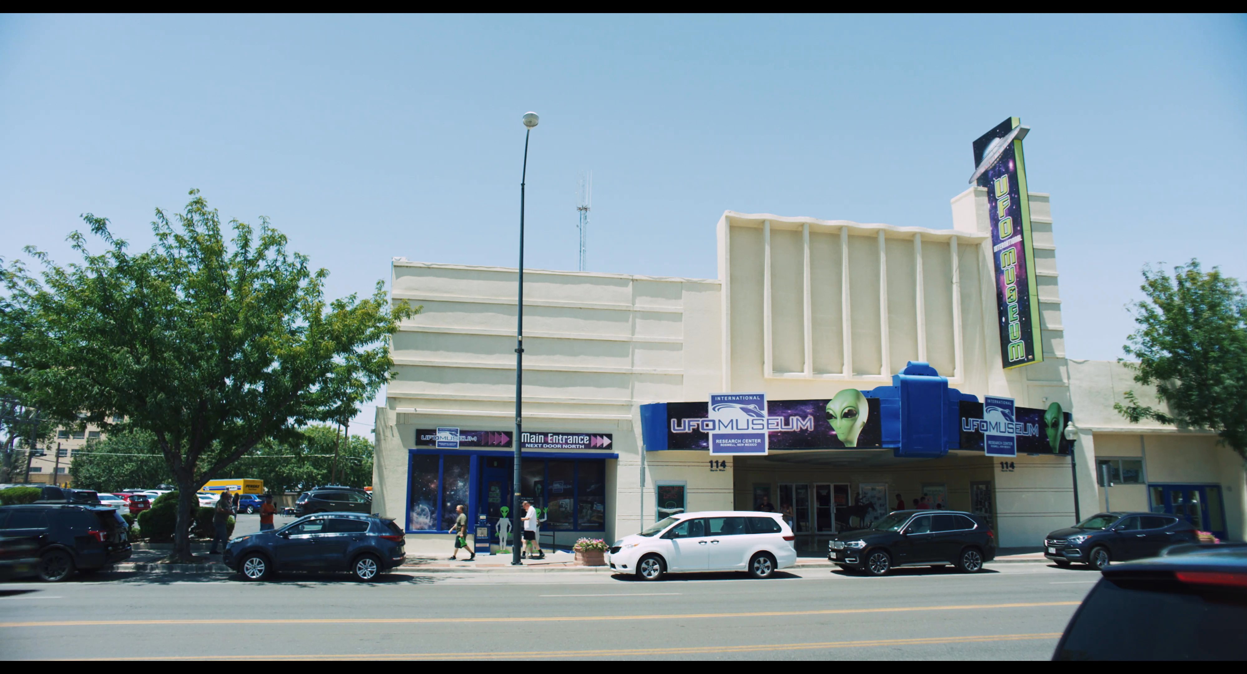 A customer browses merchandise in a roadside gift shop before transitioning to external shots of a museum building and an expansive desert road. The sequence captures the transition from a retail interaction to the journey through a remote landscape.