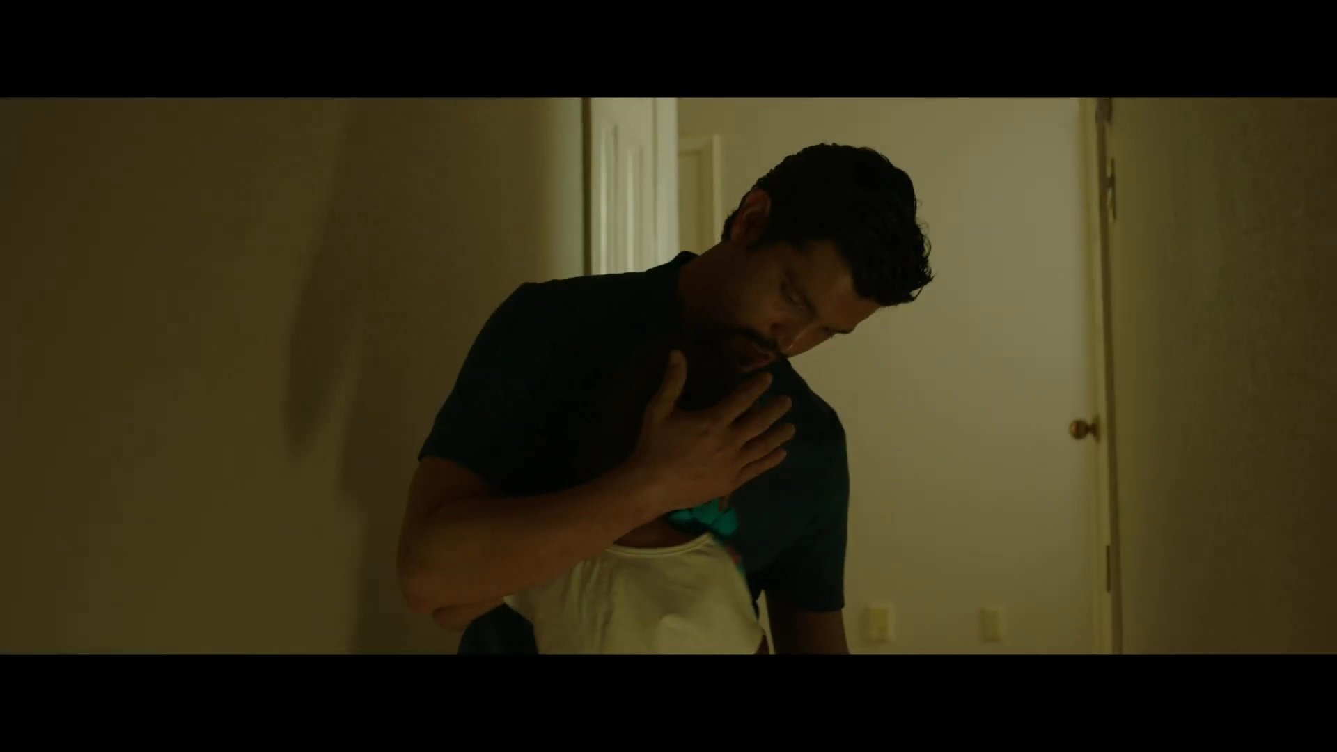 A man pauses his kitchen tasks to greet his young daughter as she runs to embrace him. He offers her a tender and protective moment, reflecting a warm familial connection.