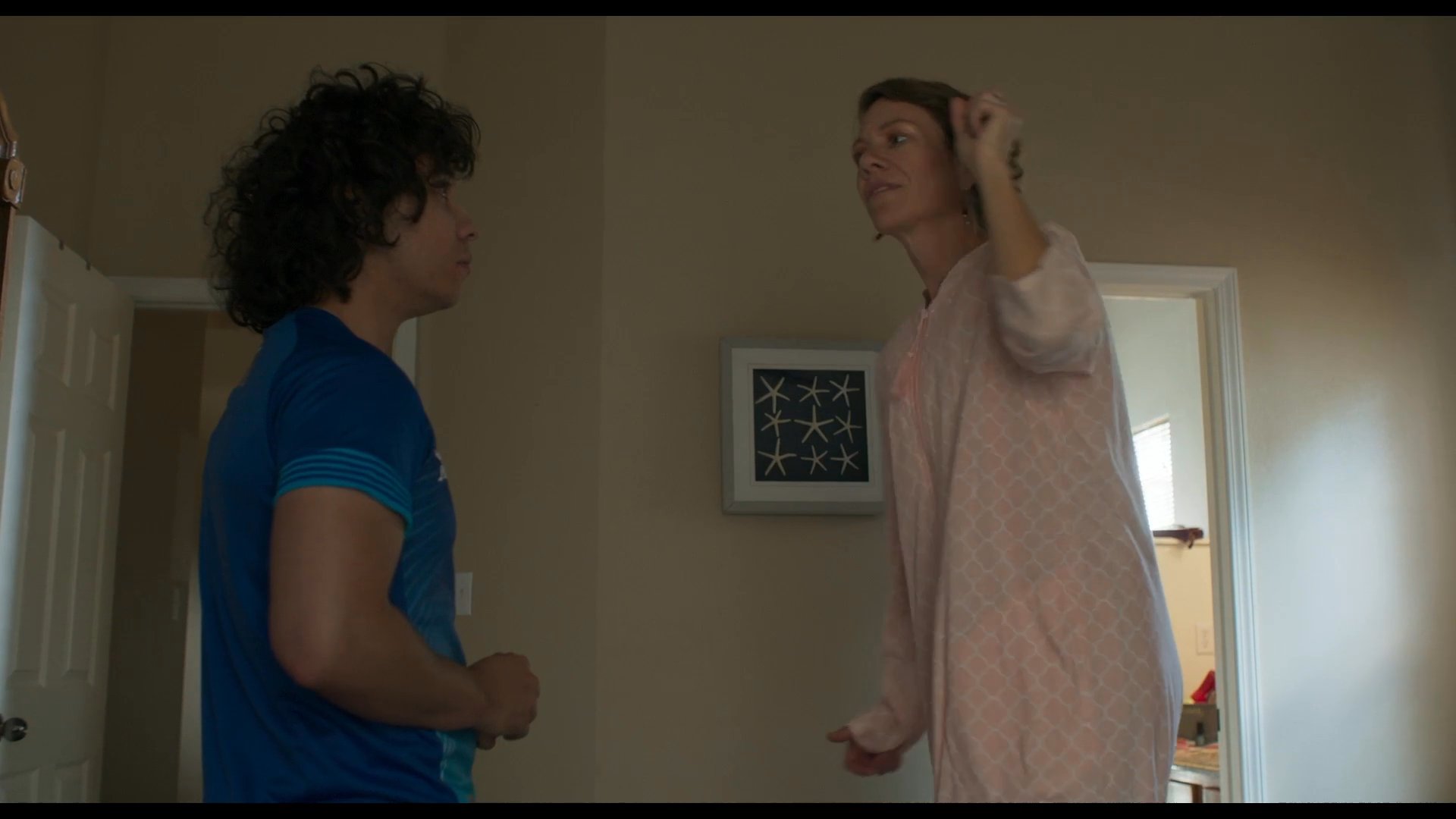 A woman in a pink robe engages in conversation with a man in an athletic shirt within a bedroom setting, eventually sharing an affectionate moment.