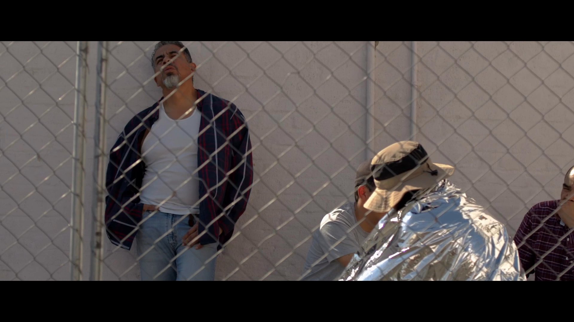 Law enforcement officers monitor a group of individuals held behind a chain-link fence. The detainees display signs of physical and emotional distress while waiting under supervision.