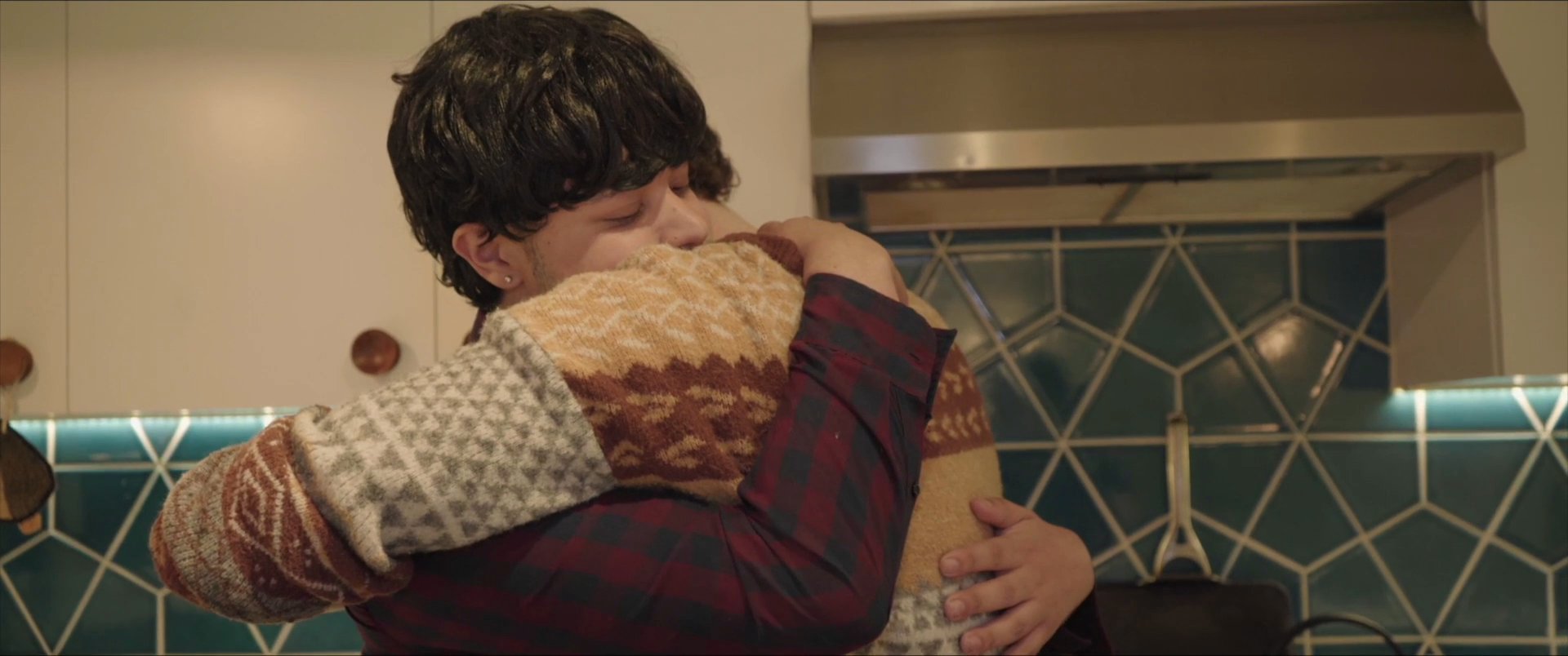 A young man speaks directly to the camera before transitioning to an intimate moment of comforting another person with a warm embrace. This sequence captures a shift from verbal communication to non-verbal physical support in a domestic setting.