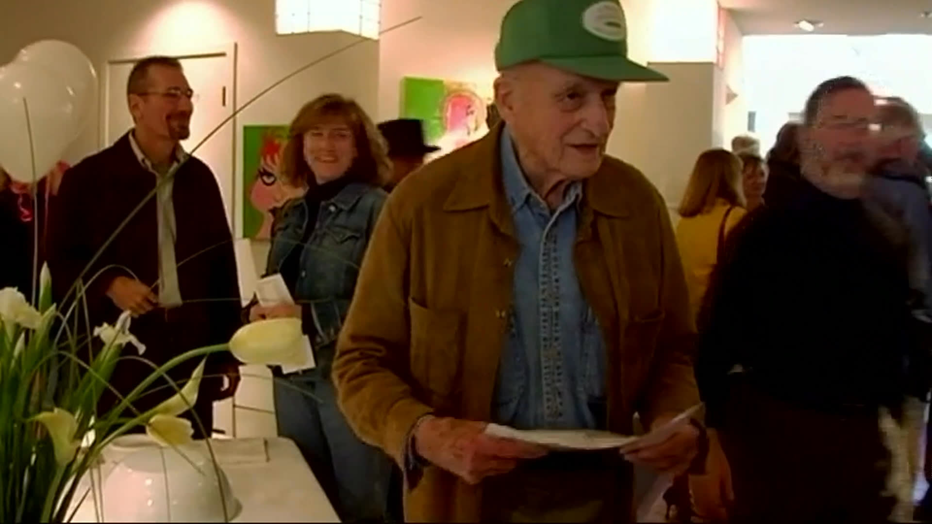 An older man in a green cap and brown jacket moves through a crowded art gallery, gesturing toward displayed works and holding printed papers. He speaks with a woman during the exhibition gathering.