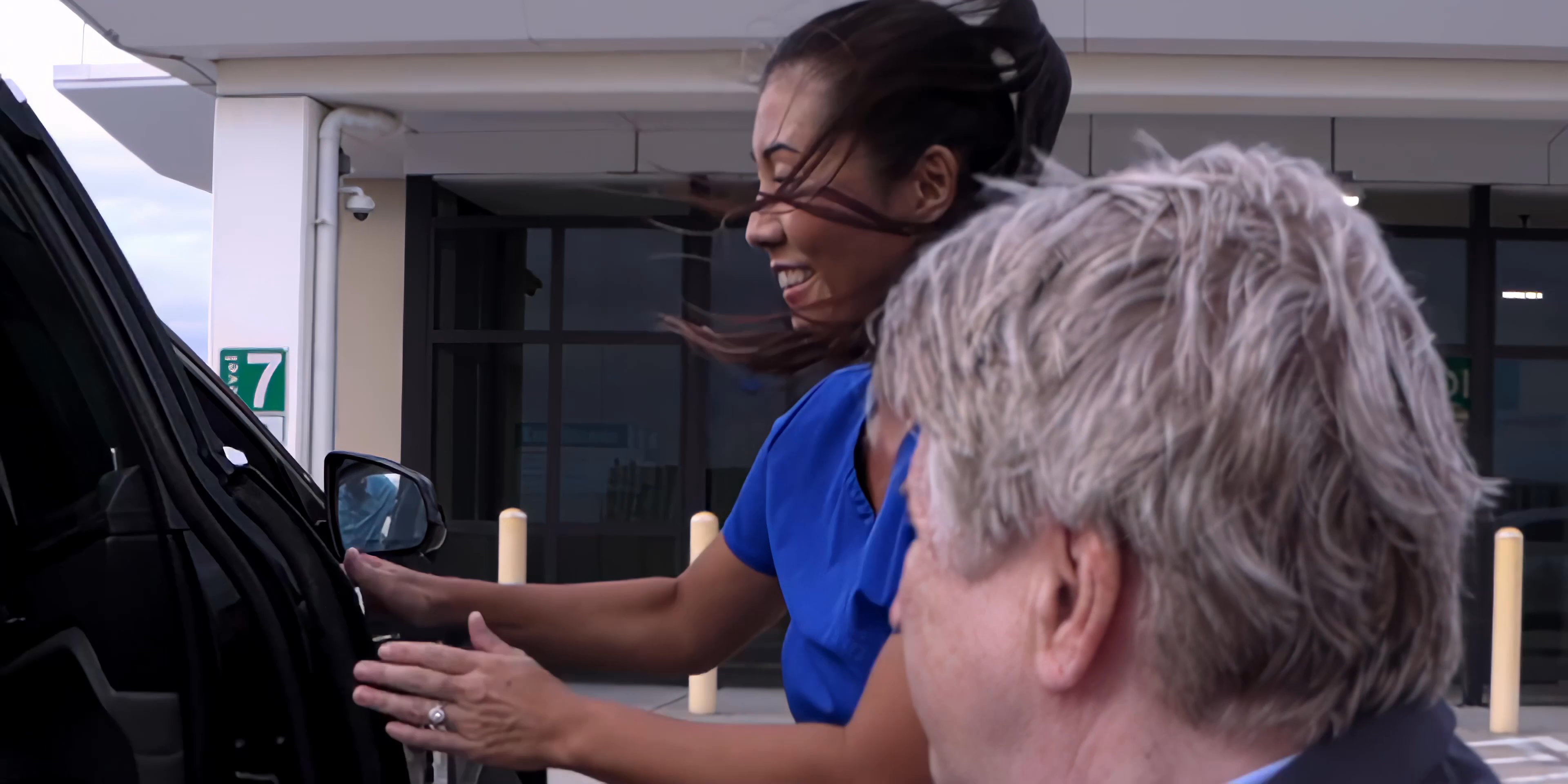A caregiver in blue scrubs assists a patient in a suit into a car parked on a hospital rooftop level. They coordinate the movement from a wheelchair into the passenger seat.