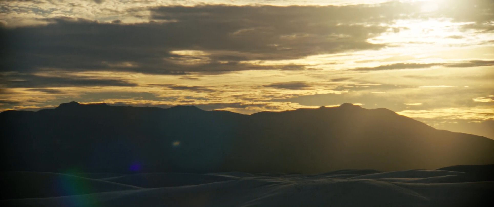 A man kneels on white sand dunes during a golden hour sunset, transitioning into a reflective, peaceful landscape composition. The scene captures a moment of quiet contemplation within a vast, arid environment.
