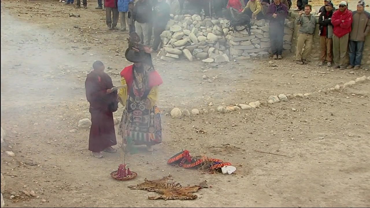 Performers in traditional ornate masks and robes enact a ceremonial ritual before an audience of spectators and monks in a mountainous region. The dance incorporates rhythmic movement and incense smoke, signifying a sacred cultural tradition.
