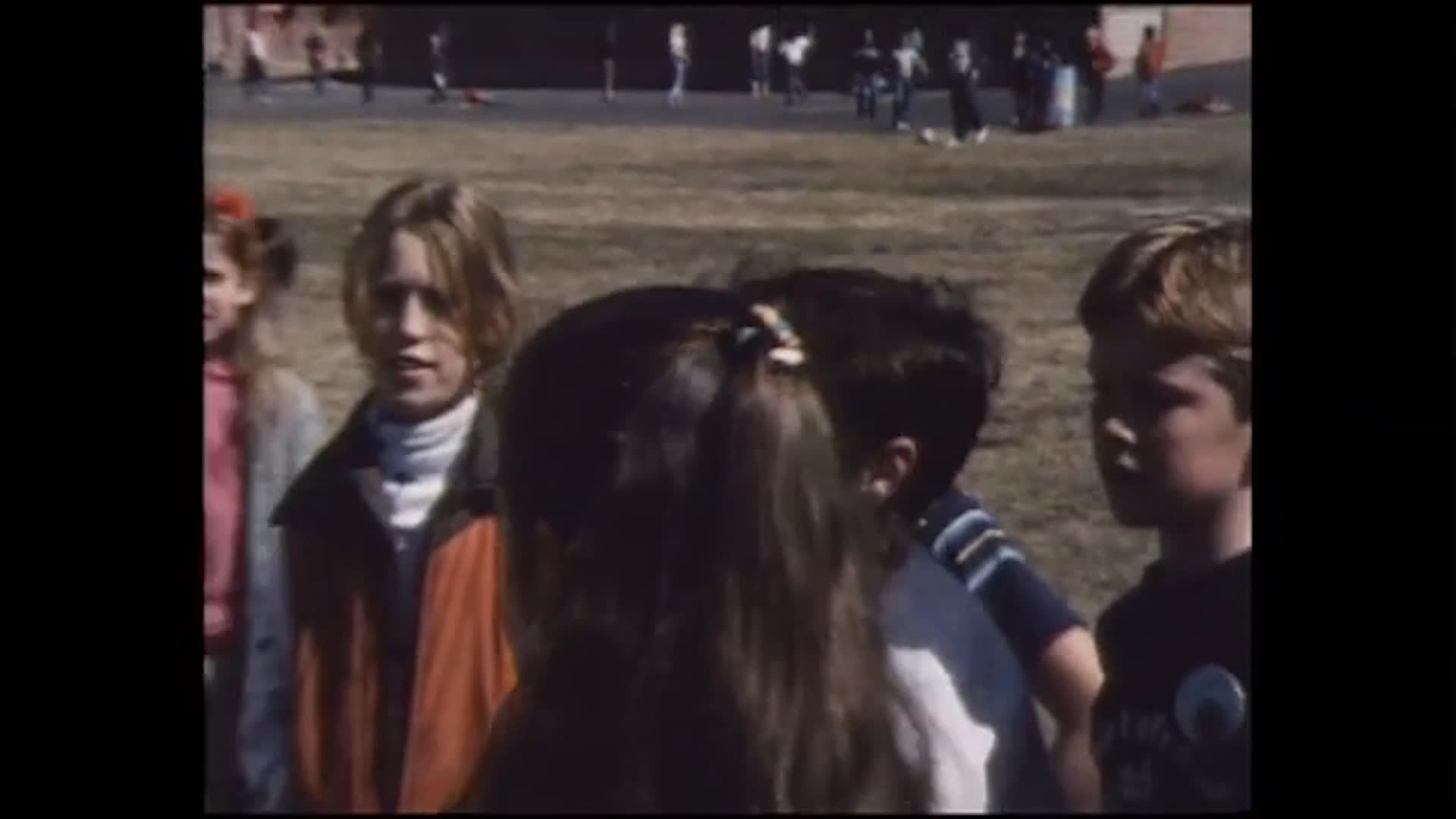 Children gather in an open field and take turns swinging a yellow tetherball around a pole as their peers watch. Earlier, a girl looks through an illustrated booklet before gazing out at a snowy rural landscape; the dialogue mentions a cowboy hat and a snowman.