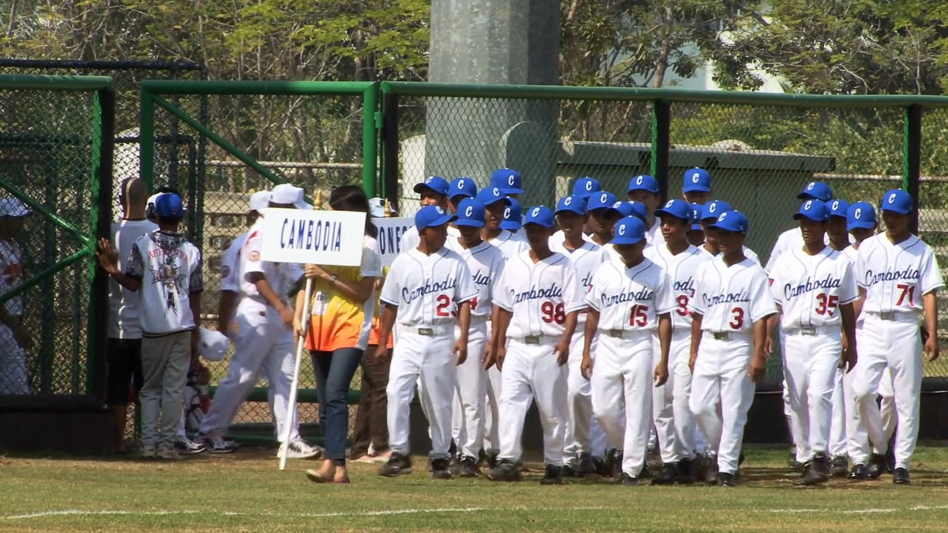 The Cambodian baseball team marches across a field during an opening ceremony procession while carrying a national placard. The athletes wear white uniforms and blue caps as they traverse the stadium ground.