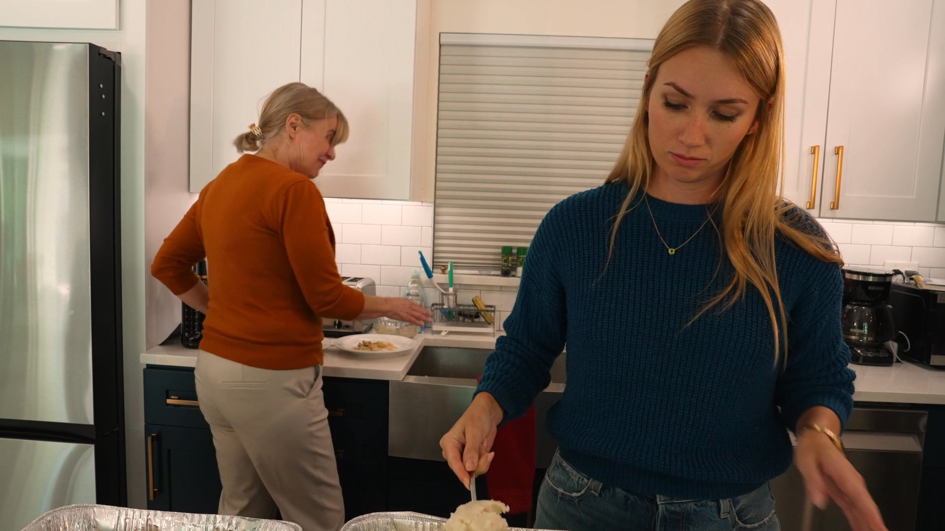 Two women prepare food in a kitchen while engaging in conversation, joined later by a man who assists with the meal service.