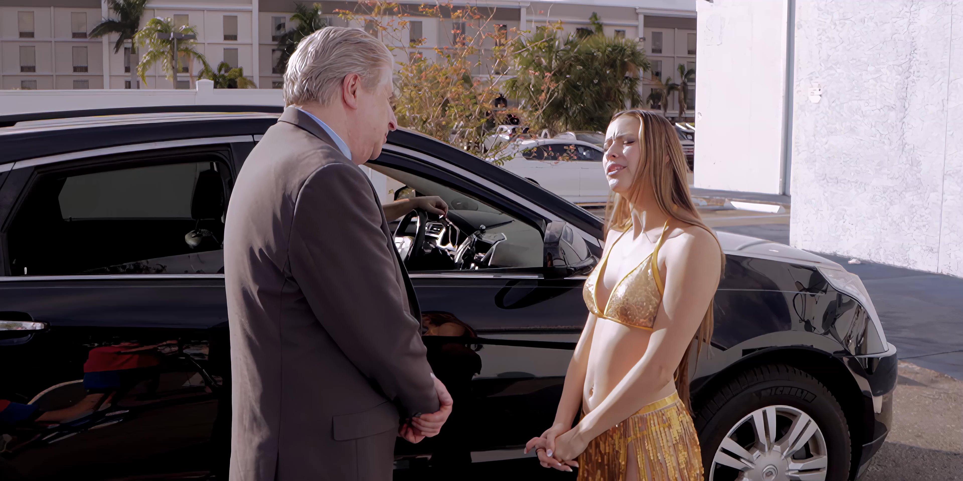 A young woman wearing a bikini and fringe skirt walks through a parking lot to reunite with an older businessman, who then engages with a man sitting in a parked SUV.