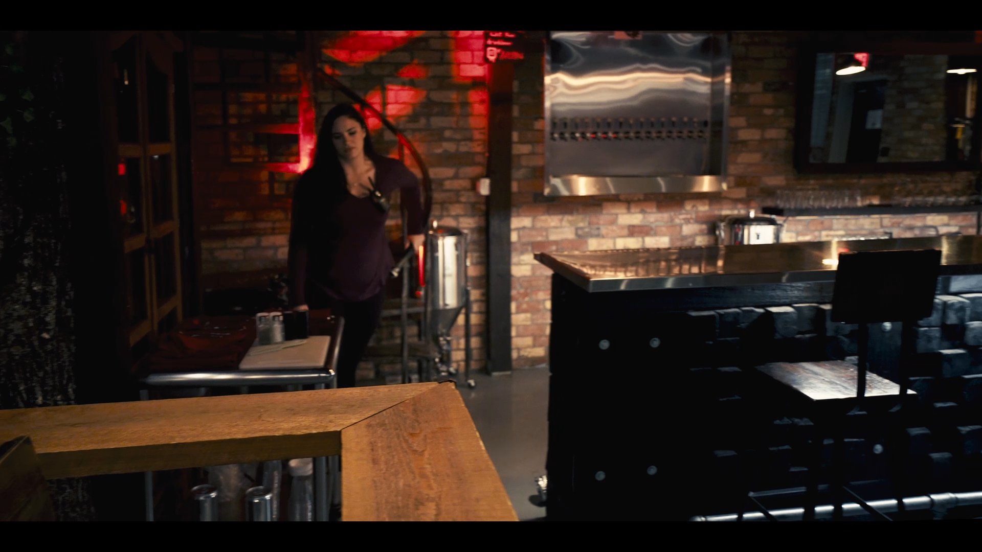 A woman and a man engage in conversation within the dimly lit interior of a modern bar. The scene transitions from a private moment near curtains to a focused discussion at the bar counter.
