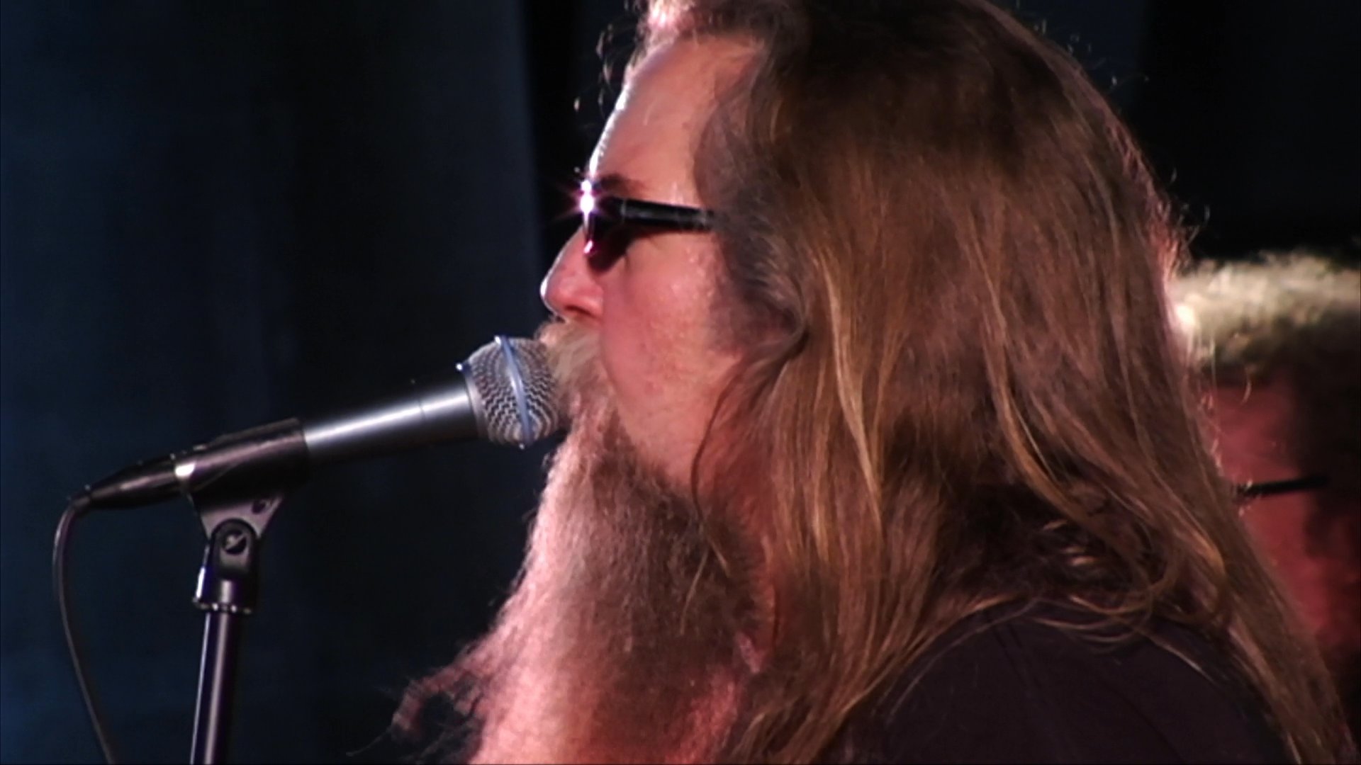 A long-haired, bearded musician performs on stage with his band, singing into a microphone during a live concert event.