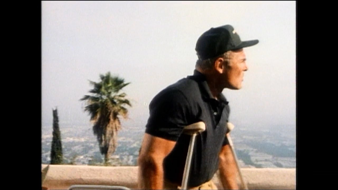 A man using crutches stands on a high-elevation balcony overlooking a city as guests move around him during a lively social gathering. He surveys the scene while interacting with attendees near a decorative gate.