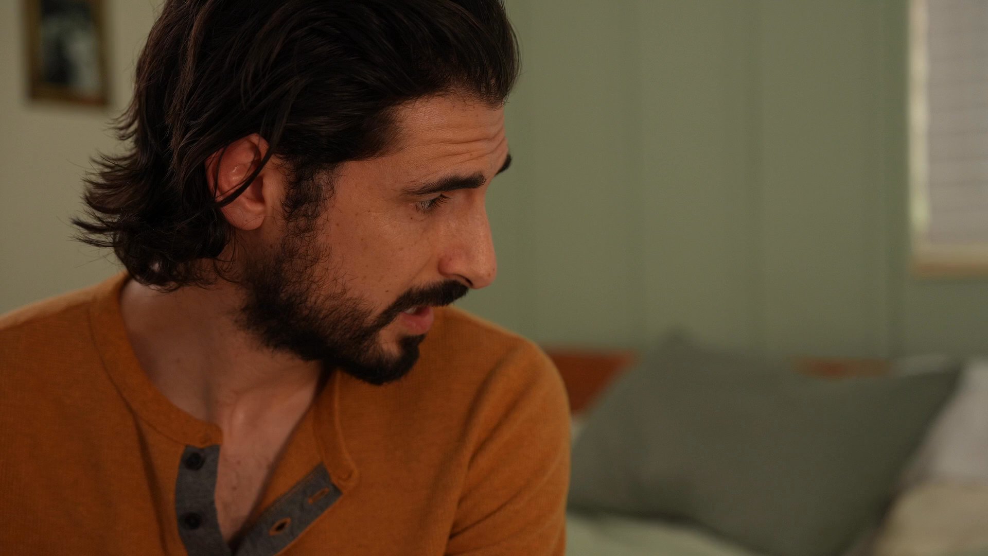 A man sits quietly in a bedroom, watching over a woman as she rests peacefully on the bed. His expression reflects a sense of concern and attentiveness toward her.