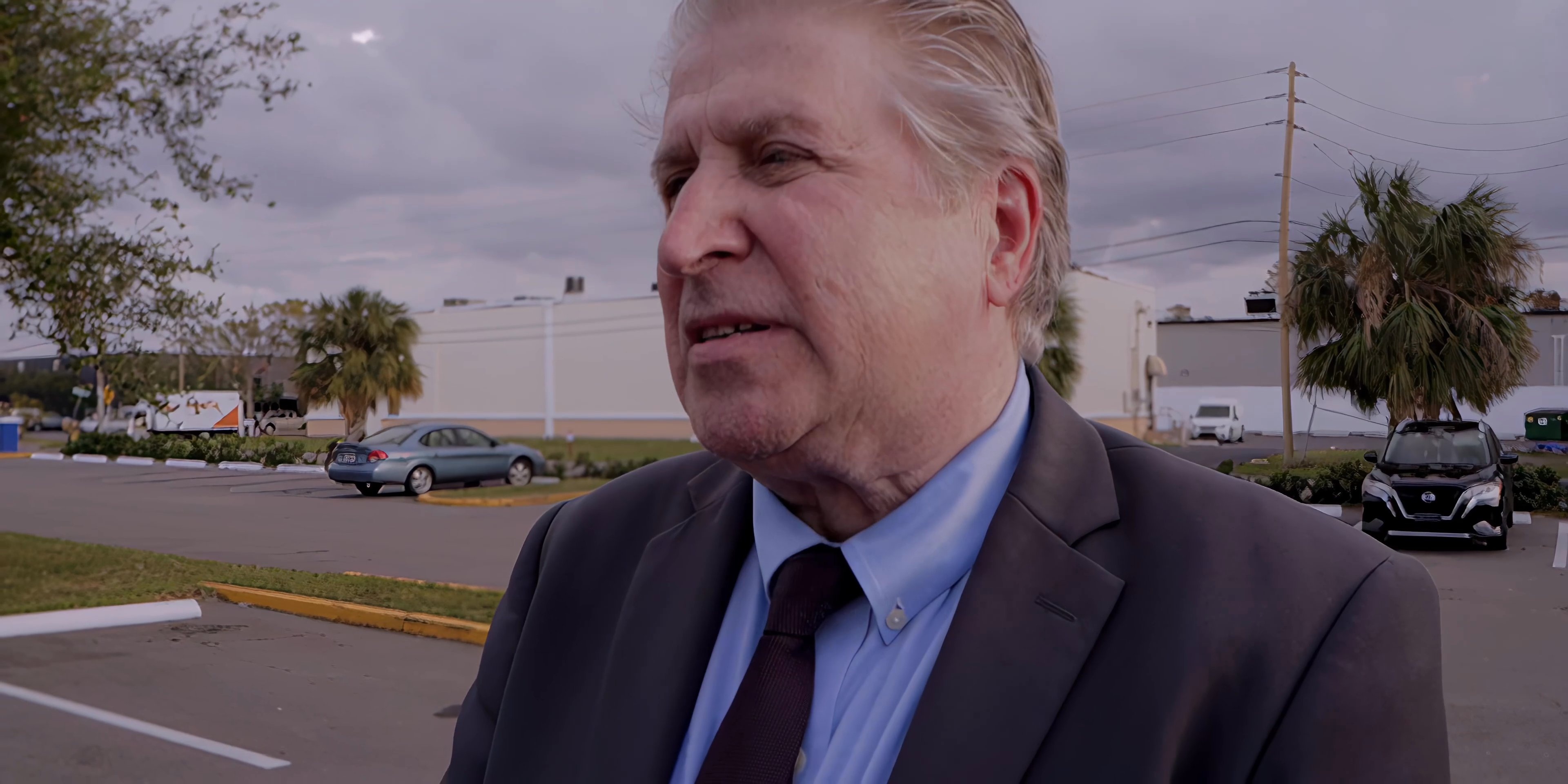 An older man in a business suit and a younger woman in an evening dress engage in a tense conversation in a parking lot. They exchange dialogue while standing near an open vehicle, displaying visible emotional strain.