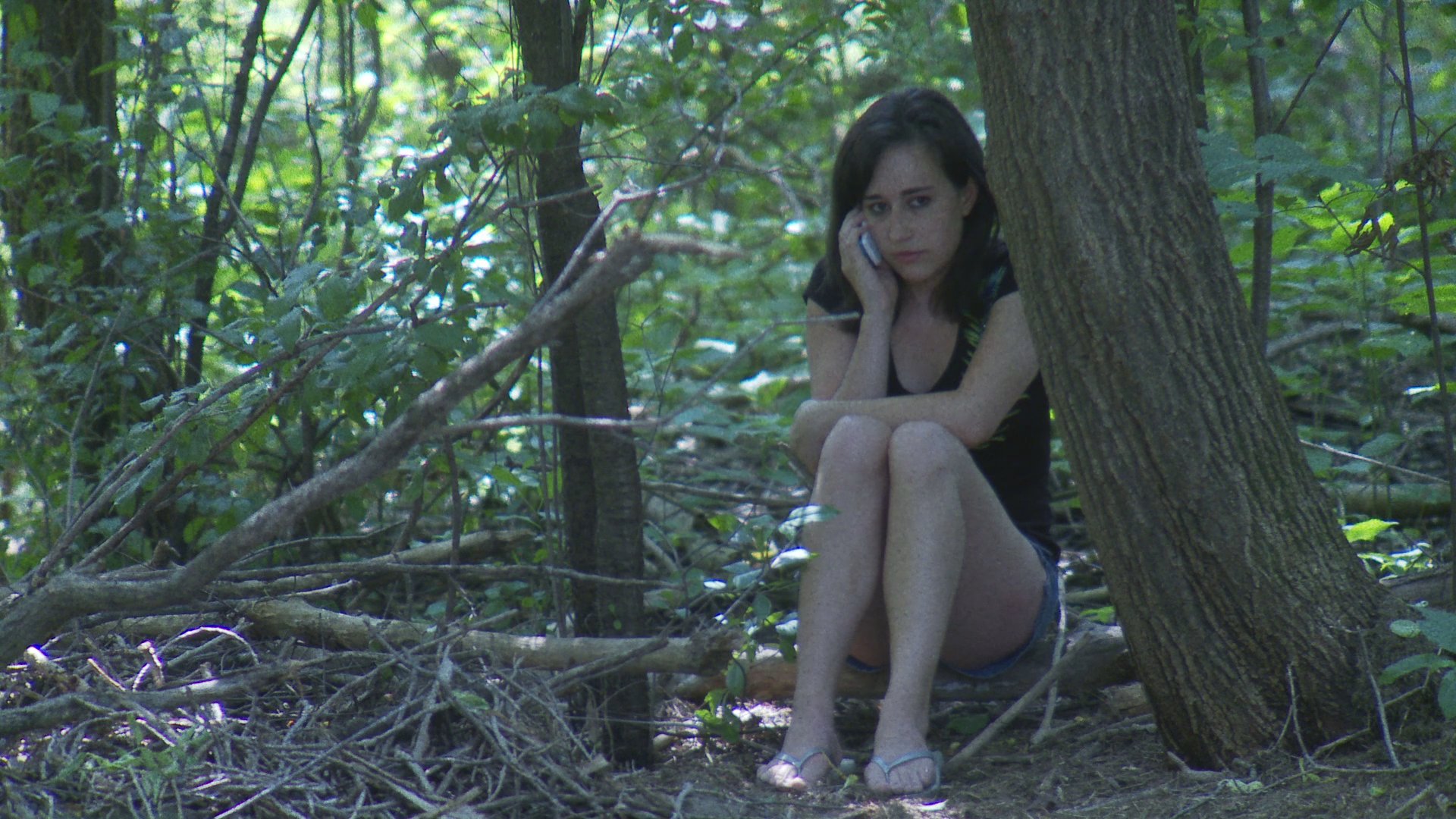 A woman sits in a woodland area while talking on her mobile phone, appearing distressed. Shortly after, another individual walks a dog nearby through the forest trees.