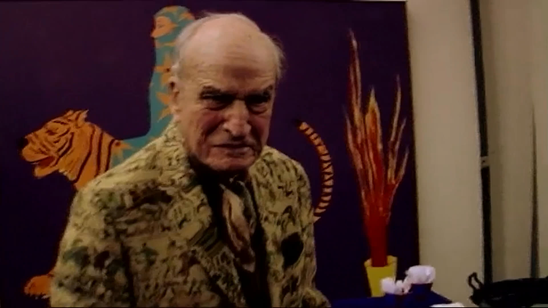 An elderly man in a distinctive patterned suit walks through a gallery as a woman recounts her surprise when he seriously asks to buy the house she made. Her story conveys a sense of shared boldness and delight at the unexpected connection.