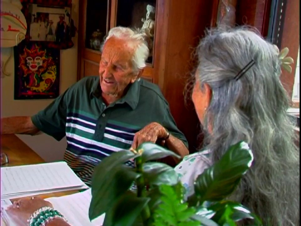 An elderly man shares insights regarding a book titled The Gospel of Love during a thoughtful conversation with a woman in an indoor setting. The scene transitions from a serene coastal sunrise to an intimate dialogue, highlighting the reflective nature of their exchange.