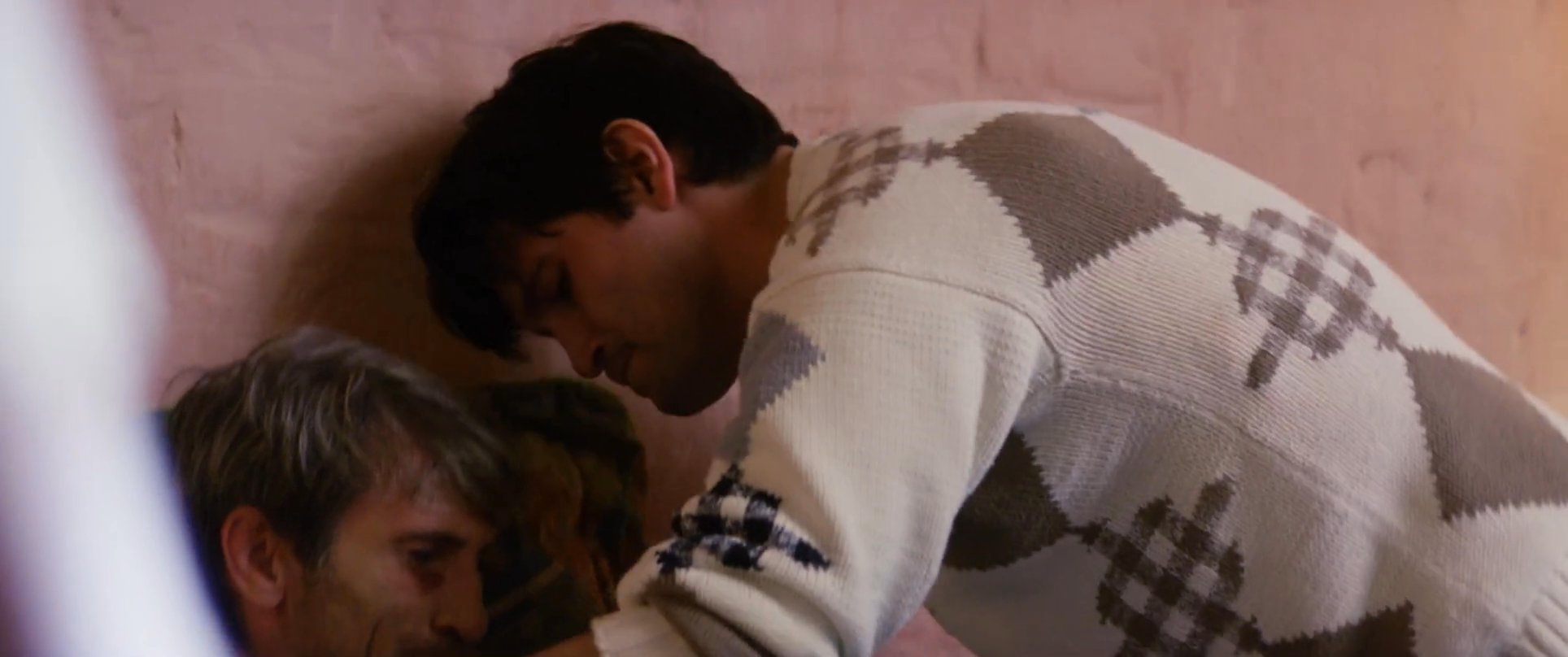 A man provides gentle physical support and comfort to an ailing individual while a woman watches nearby with concern. The sequence captures an intimate moment of caregiving and emotional connection within a home environment.
