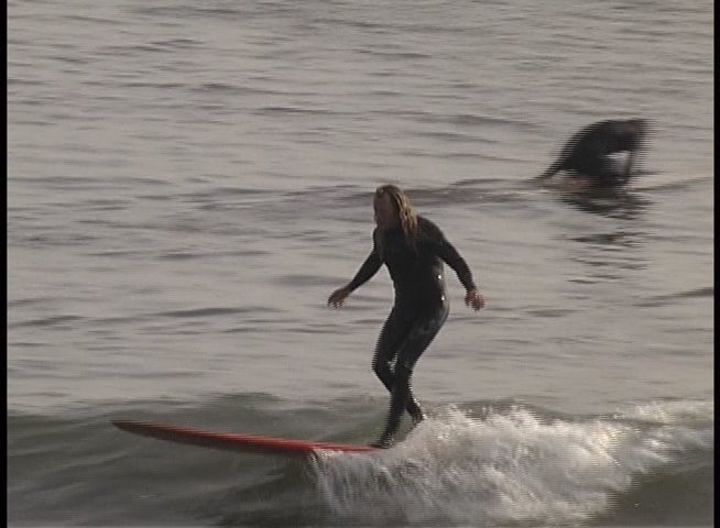 A surfer rides waves along the coastline before transitioning to an interview with an older man outdoors near a brick building.