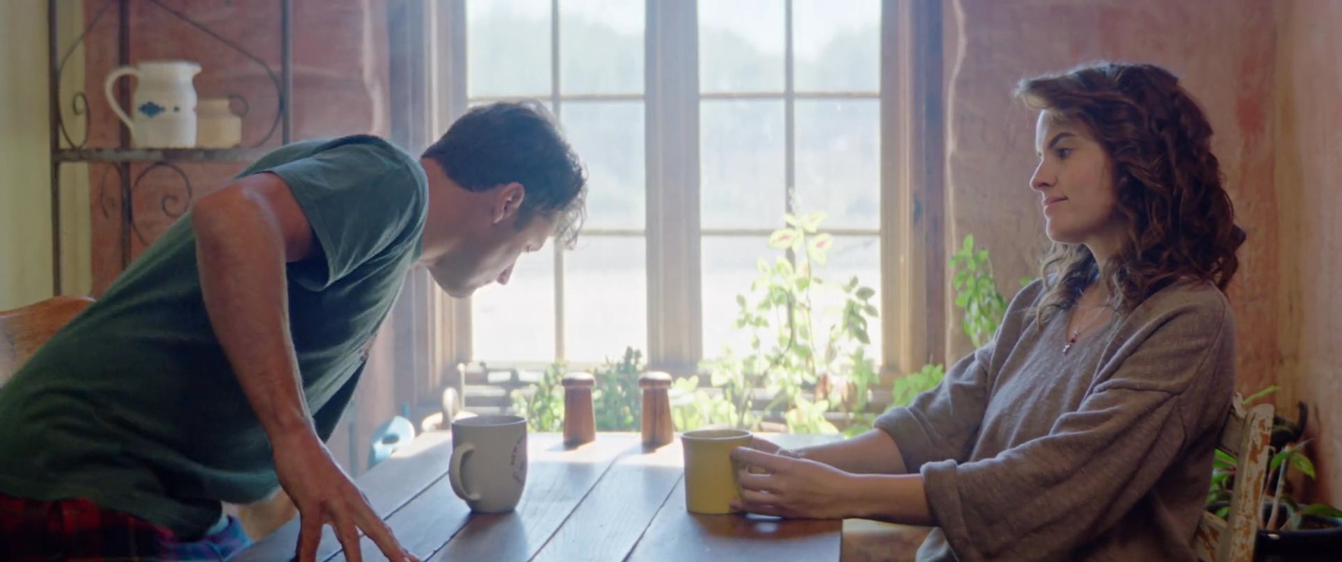 A man wakes up and joins his partner at a kitchen table for a quiet morning discussion. They share a moment of connection while sitting near a window with natural light.