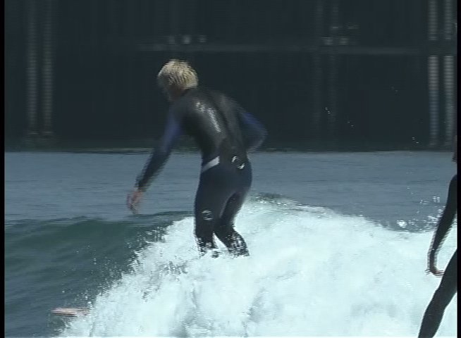 A surfer in a wetsuit maneuvers a surfboard across incoming ocean waves near a pier. The athlete maintains balance and navigates the break in a coastal setting.