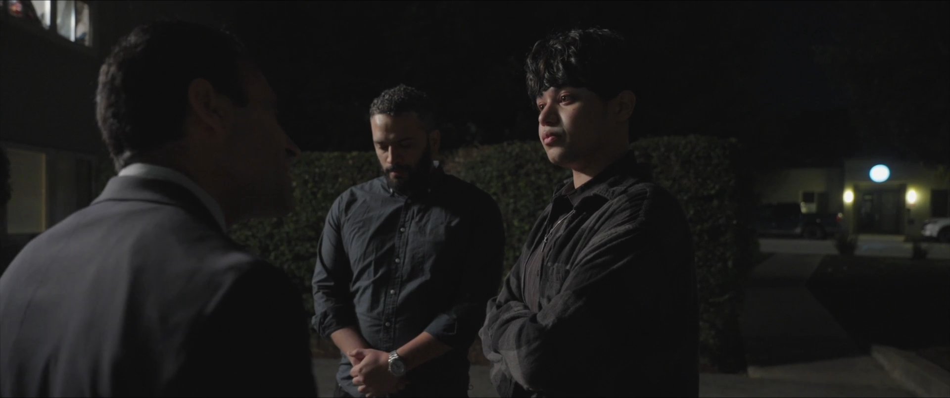 Three men engage in a serious, tense conversation in a dimly lit parking lot at night. Later, one of the men sits alone in a bedroom, lost in contemplation.