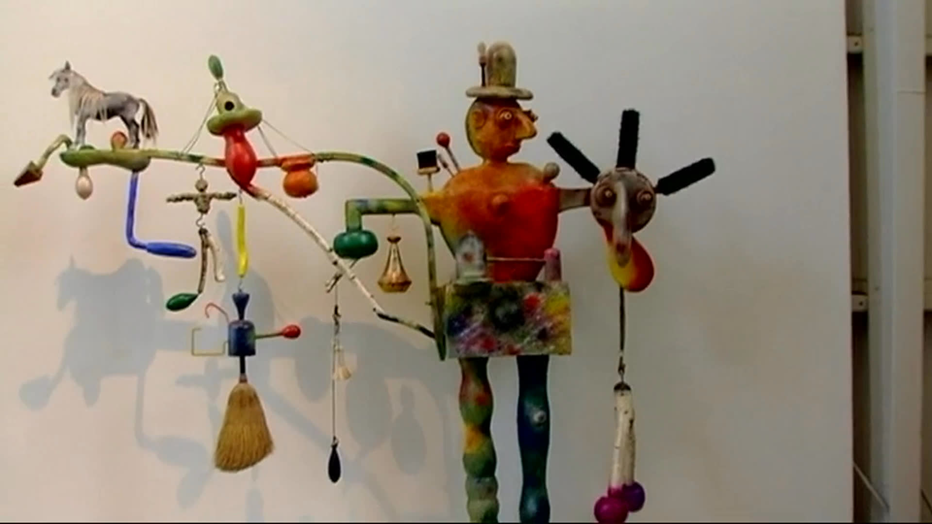 An elderly artist and a gray-haired visitor discuss a large, whimsical assemblage sculpture in a gallery, alongside close views of its colorful figures and sculpted heads. The conversation turns to the artwork’s size and the artist’s traveling days.